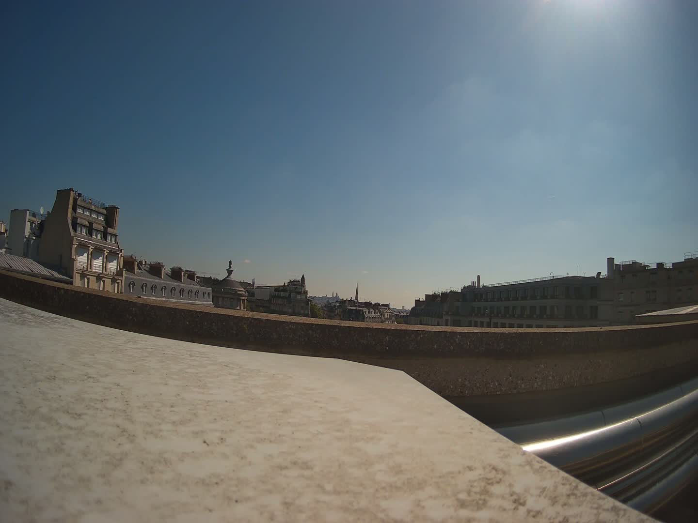 A mostly sunny day reveals a cityscape of Parisian rooftops and buildings, viewed from a low angle with a large, light-colored surface in the foreground.