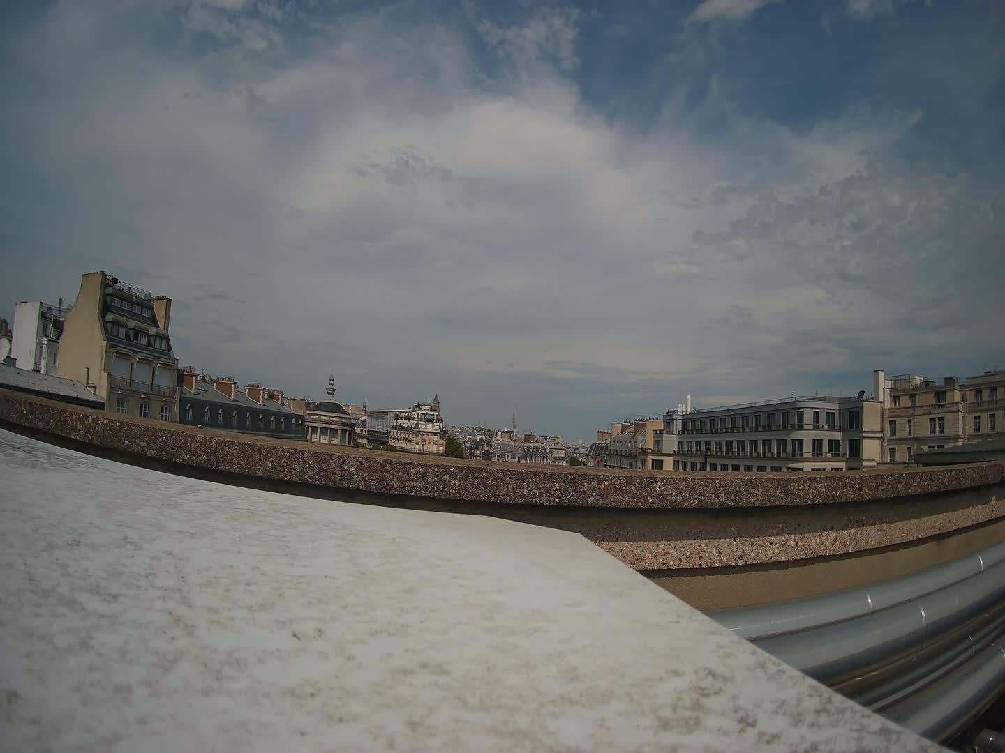 A mostly cloudy sky overlooks a cityscape of Parisian buildings viewed from a rooftop, with a large, slightly dirty white surface in the foreground.