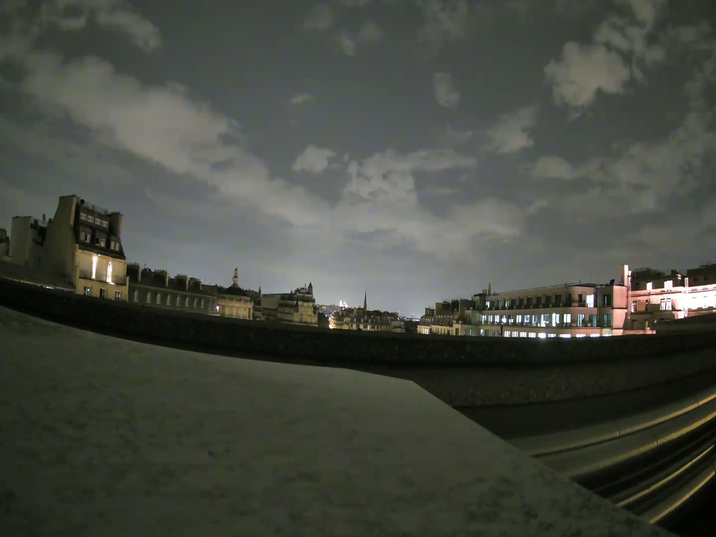 A nighttime cityscape view of Parisian buildings under a partly cloudy sky is seen from a rooftop vantage point.