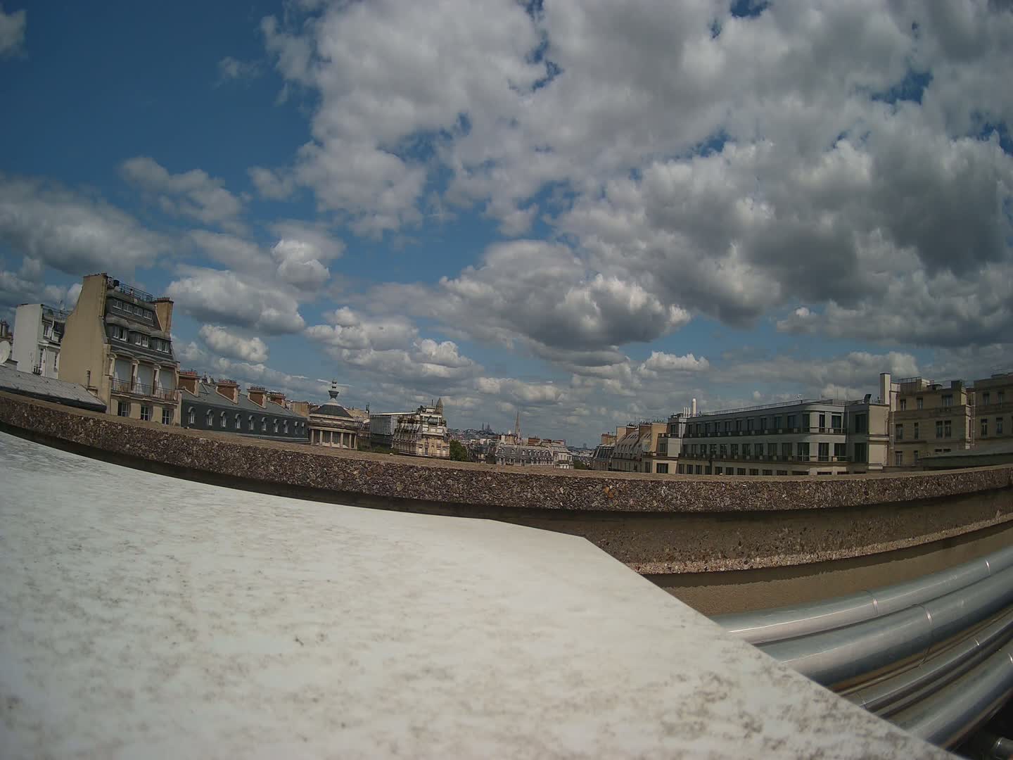 A partly cloudy sky above a cityscape of Parisian-style buildings viewed from a rooftop with large metallic pipes in the foreground.
