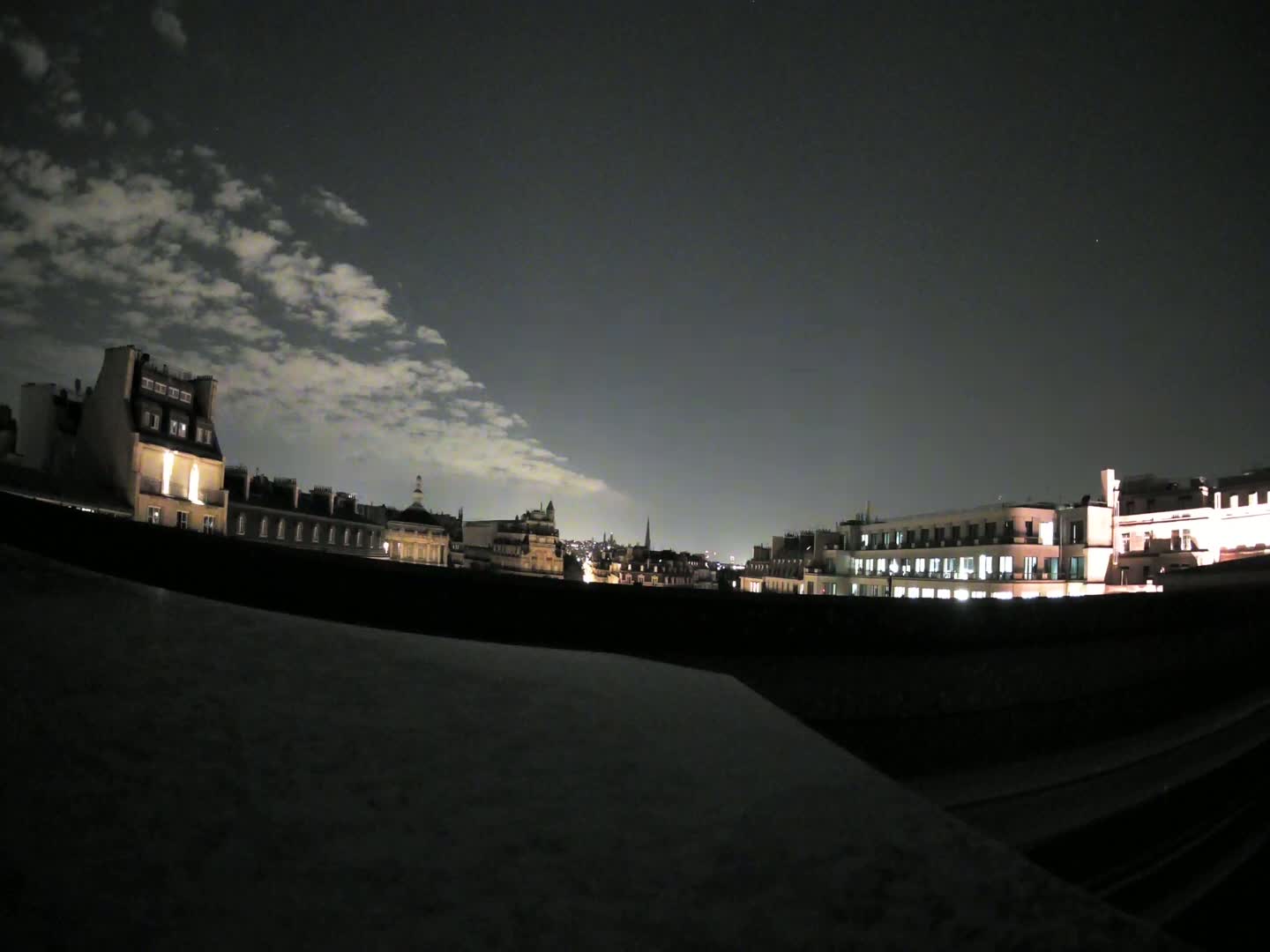 A nighttime cityscape with partly cloudy skies is seen from a rooftop vantage point.