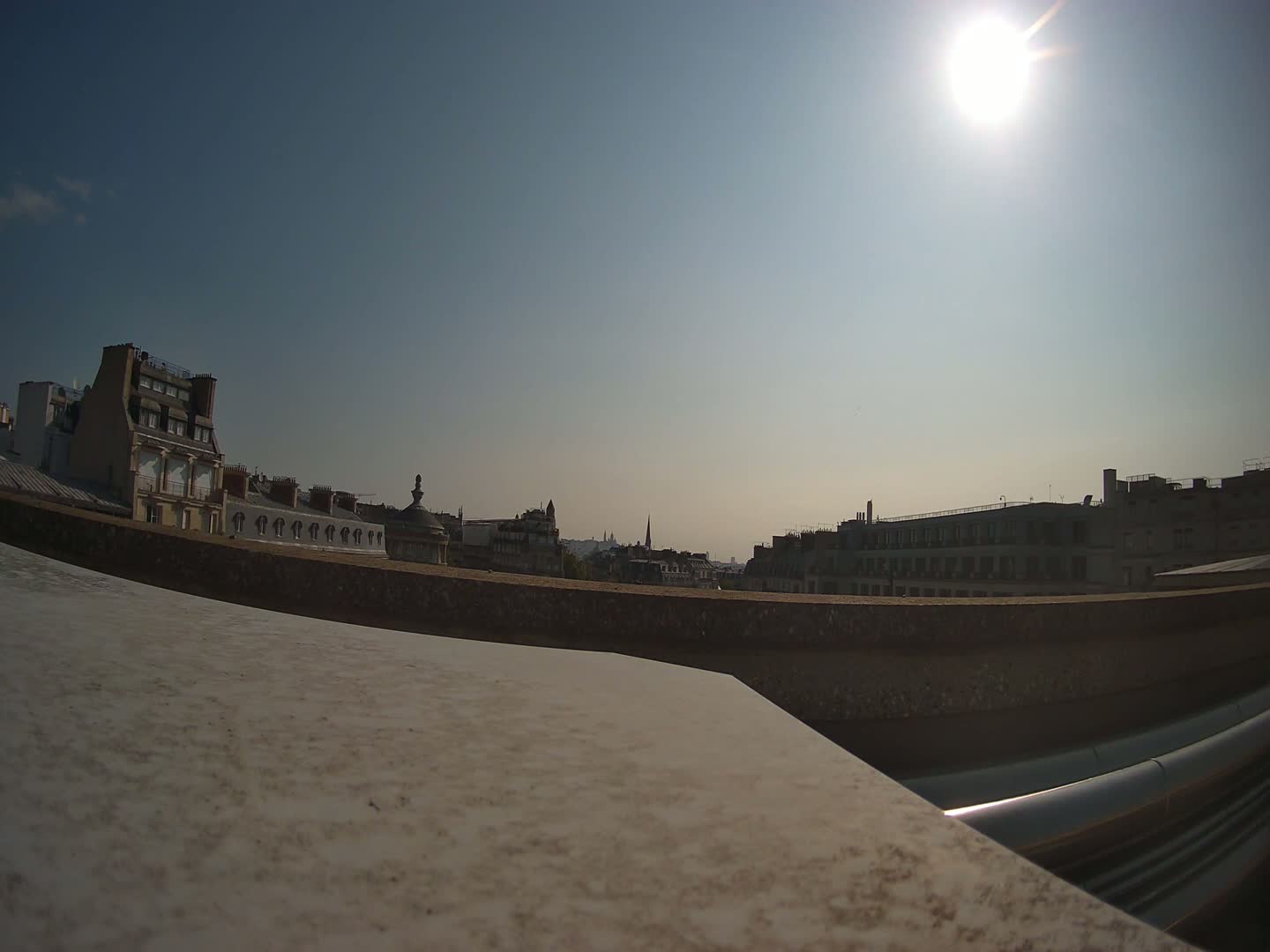A sun-drenched cityscape viewed from a rooftop, showing several multi-story buildings under a bright, clear sky.