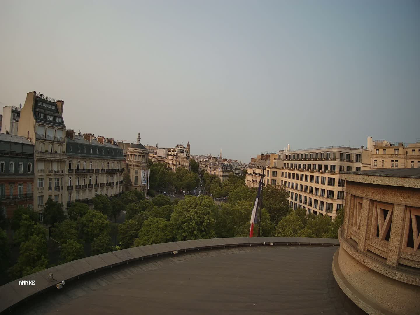 A Parisian cityscape with leafy trees lining a street, viewed from a rooftop under a clear, dusky sky.