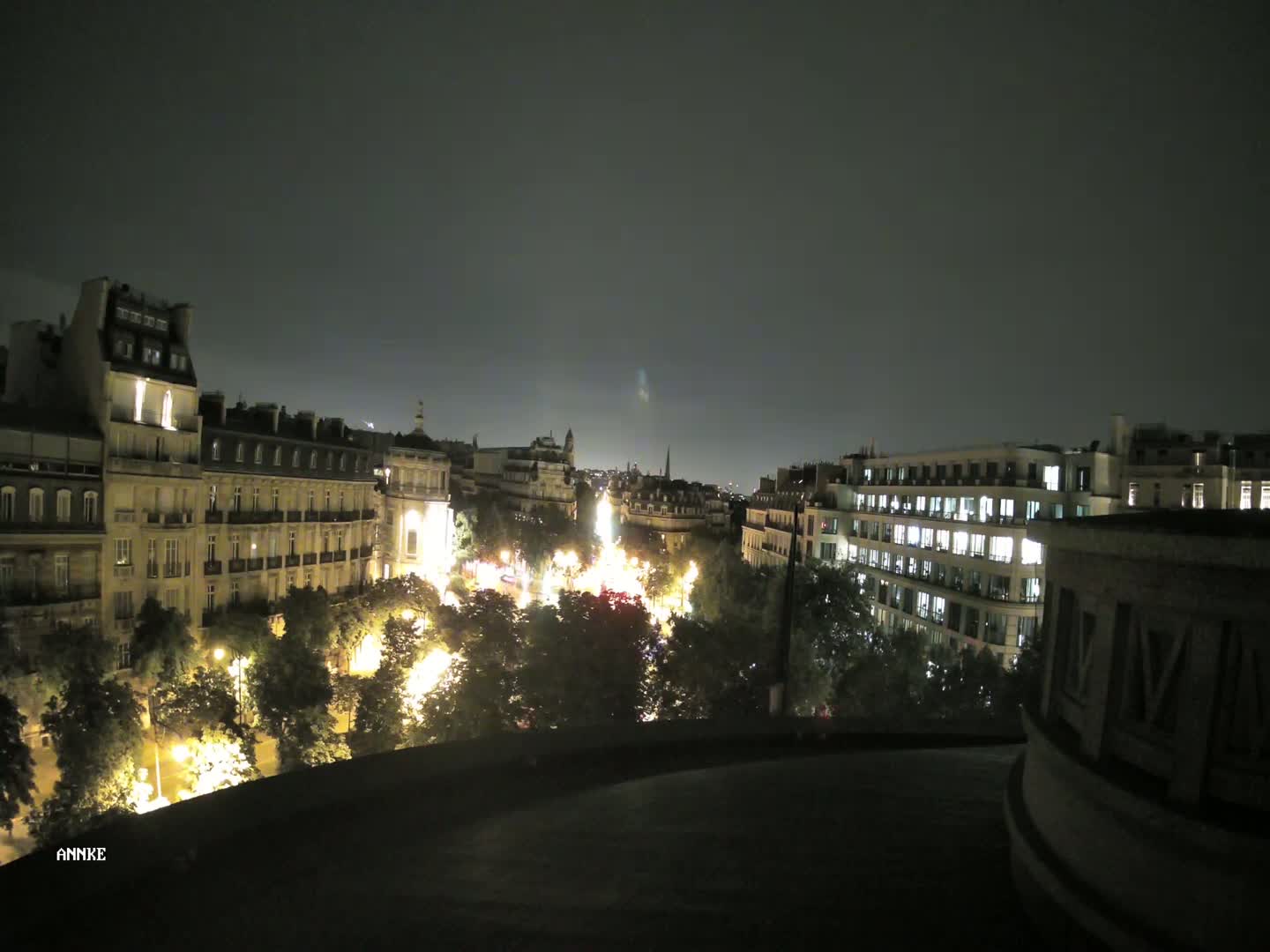 A nighttime view of a city street lined with illuminated buildings and trees under a dark sky.