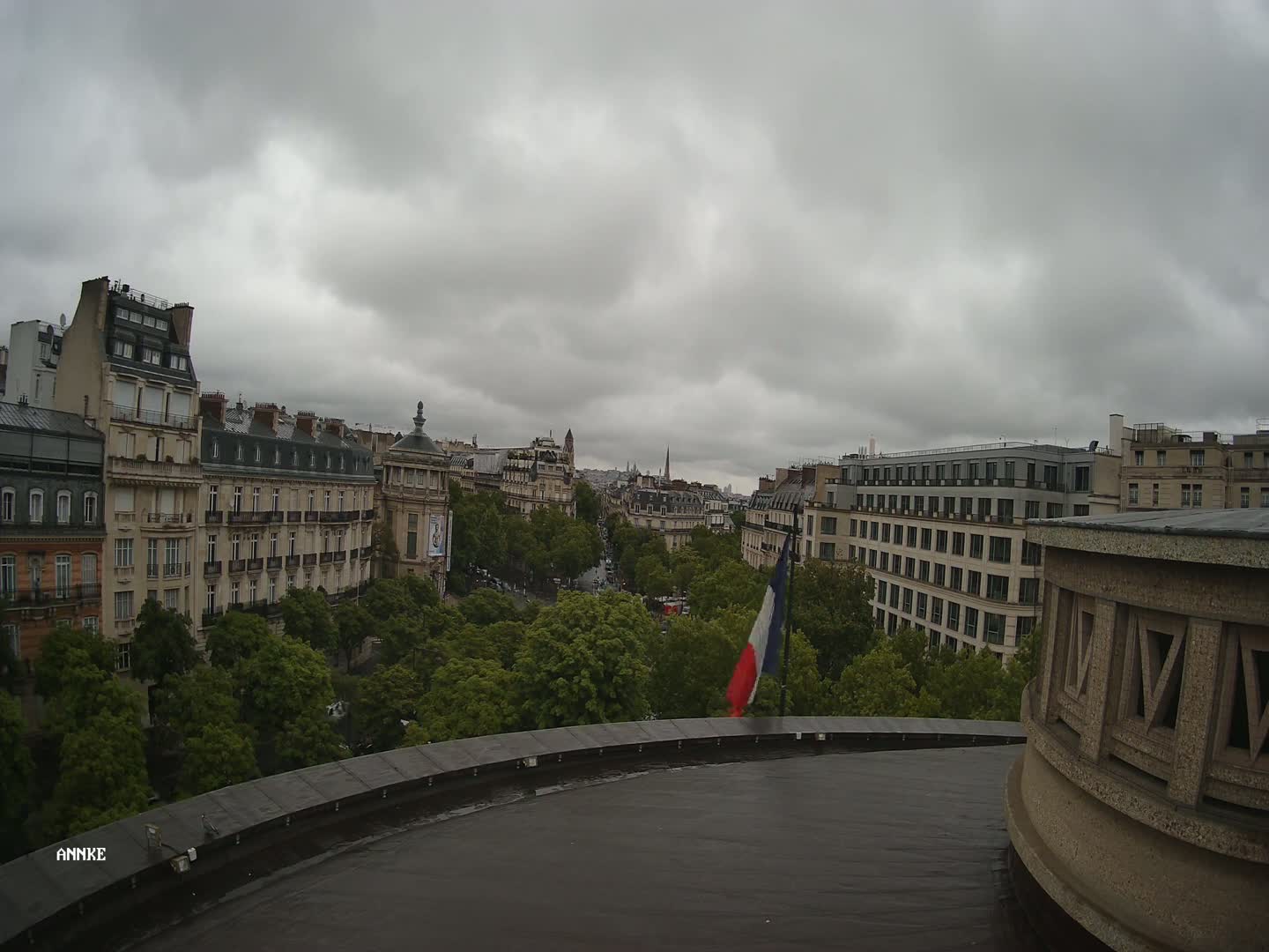 A view from a rooftop shows a city street lined with buildings under a cloudy sky.