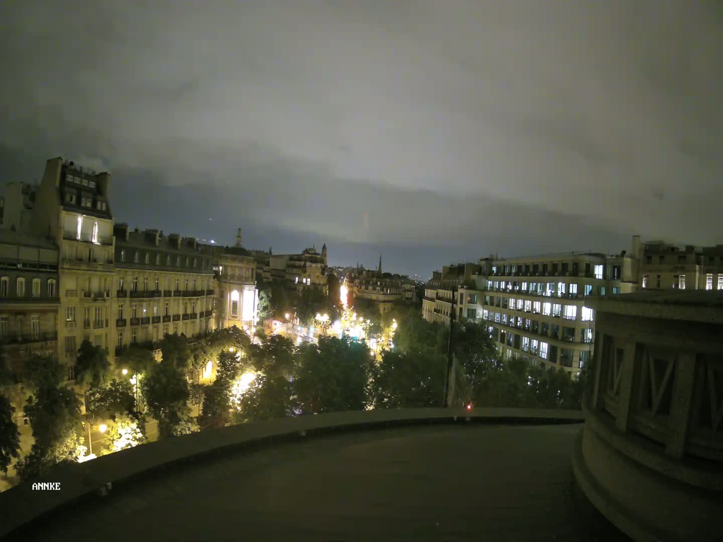 A nighttime view from a rooftop shows a city street lined with illuminated buildings and trees under a cloudy sky.