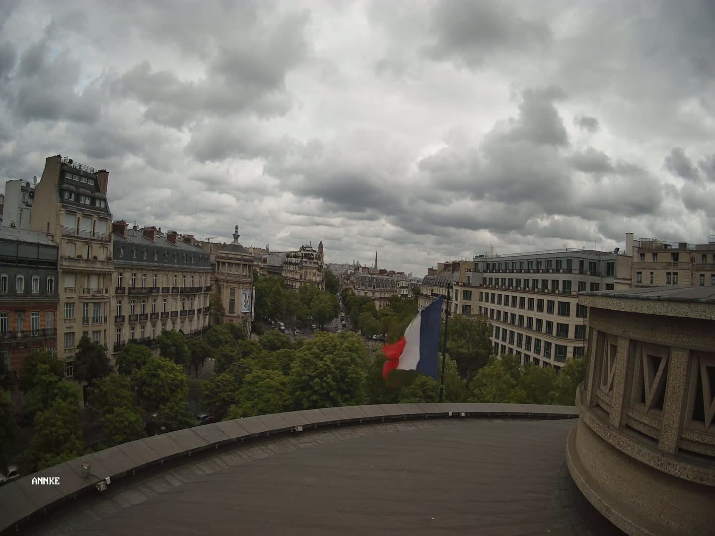 A French flag waves on a rooftop overlooking a city street lined with tall buildings under a cloudy sky.