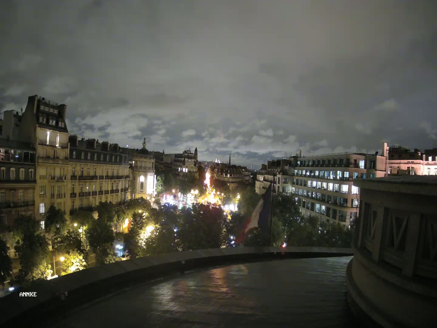 A nighttime view of a city street lined with tall buildings and trees, illuminated by streetlights under a cloudy sky.