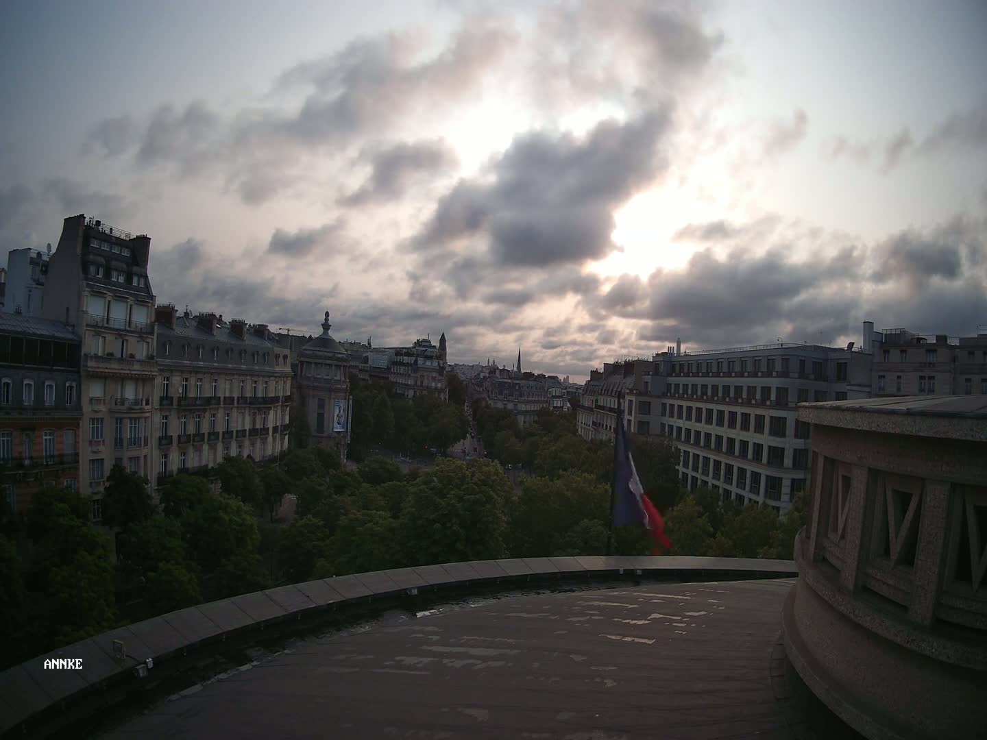 A partly cloudy sunset casts shadows over a cityscape of Parisian buildings and trees, as seen from a rooftop with a French flag in the foreground.