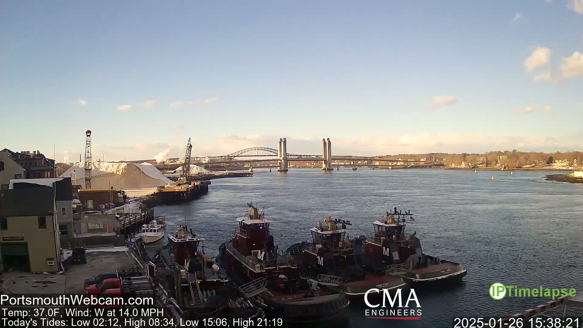 A sunny day shows a harbor with several tugboats docked, a large pile of white material, and a bridge in the distance.