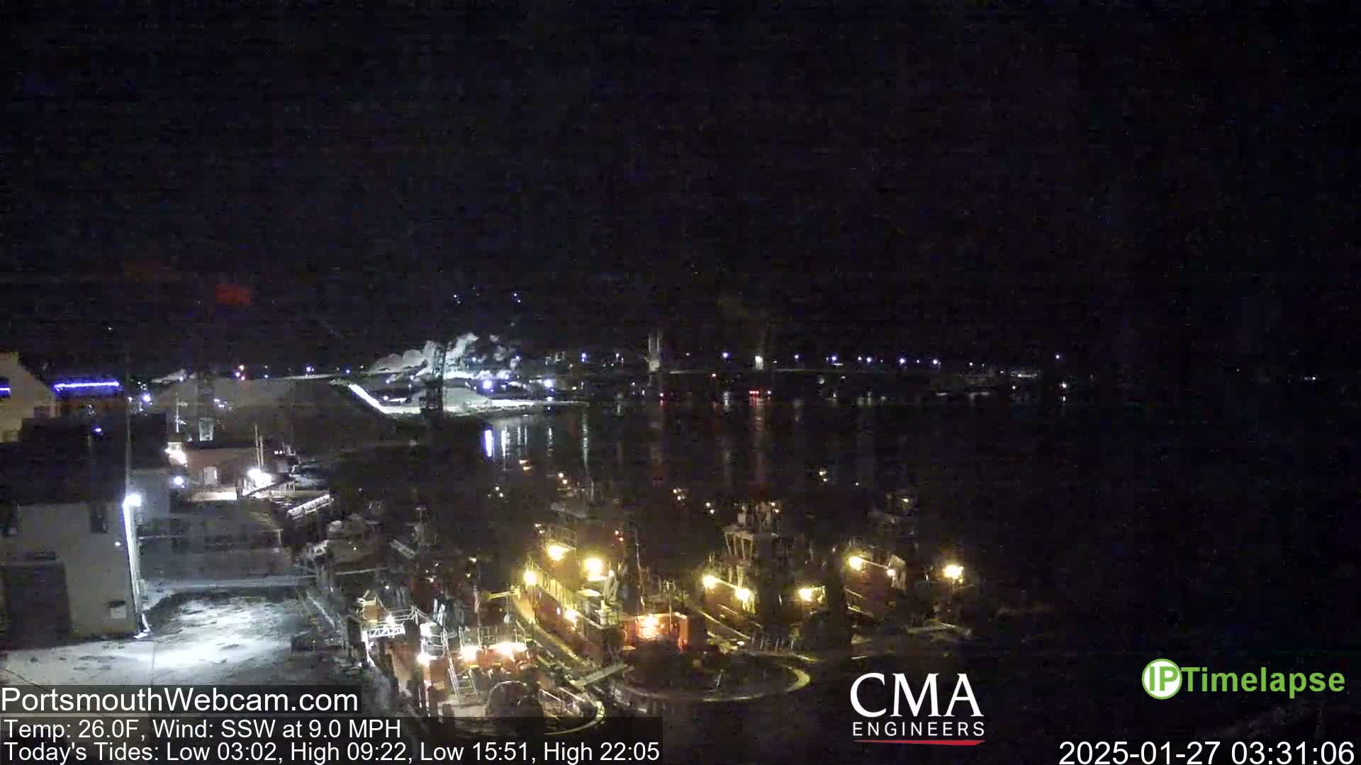 At night, under a dark sky with a south-southwesterly wind of 9 mph and a temperature of 26°F, several illuminated tugboats are moored at a dock next to a waterfront industrial area.