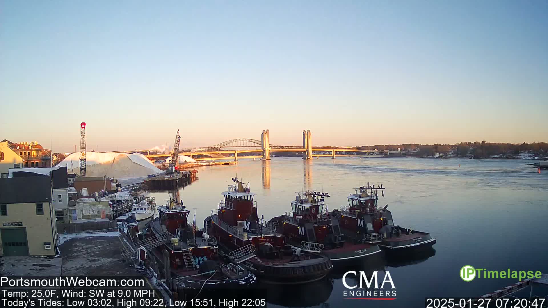 Several tugboats are moored at a dock next to a large pile of what appears to be salt, with a bridge spanning a calm river under a clear, sunrise sky.