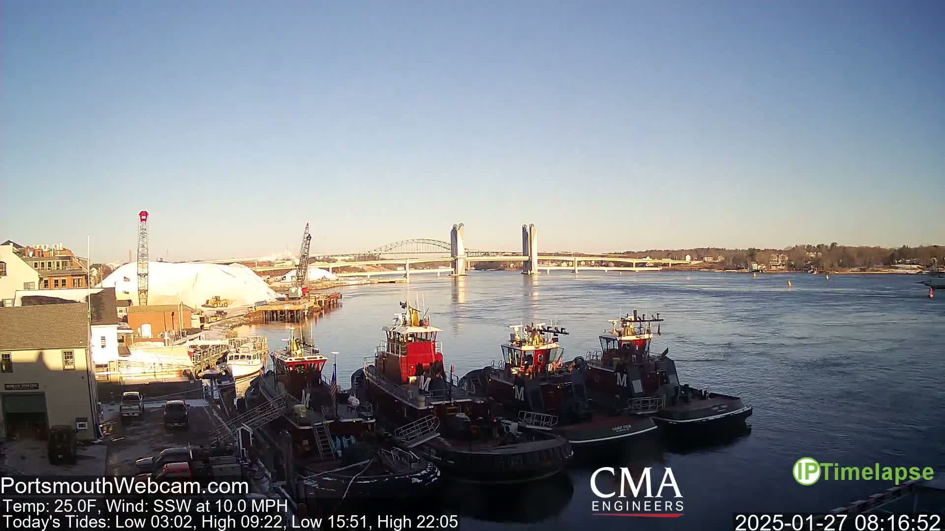 Several tugboats are moored at a dock, with a large pile of white material nearby, under a clear, sunny sky, and a bridge in the distance across a body of water.