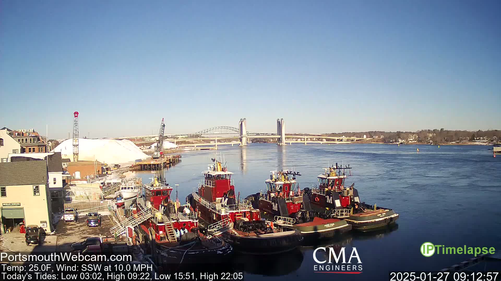 Several red tugboats are docked at a wharf next to a large pile of salt, with a bridge spanning a waterway in the background under a clear blue sky.