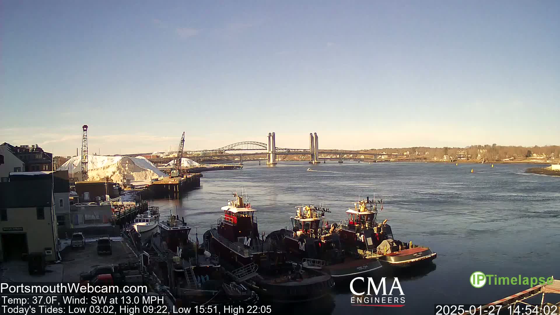 Several tugboats are moored at a dock next to a large pile of snow, with a double-leaf bascule bridge visible in the background on a sunny, breezy day.