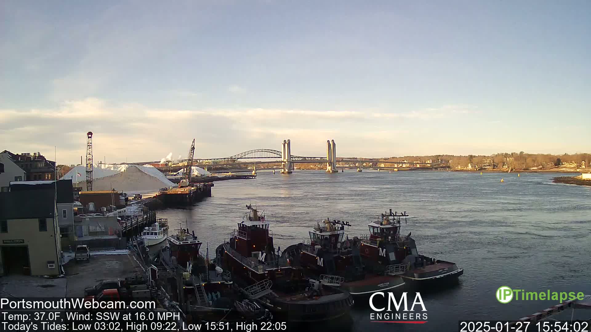 A clear, sunny day shows a harbor with several tugboats docked, a large pile of white material, and a double-bascule bridge in the distance.