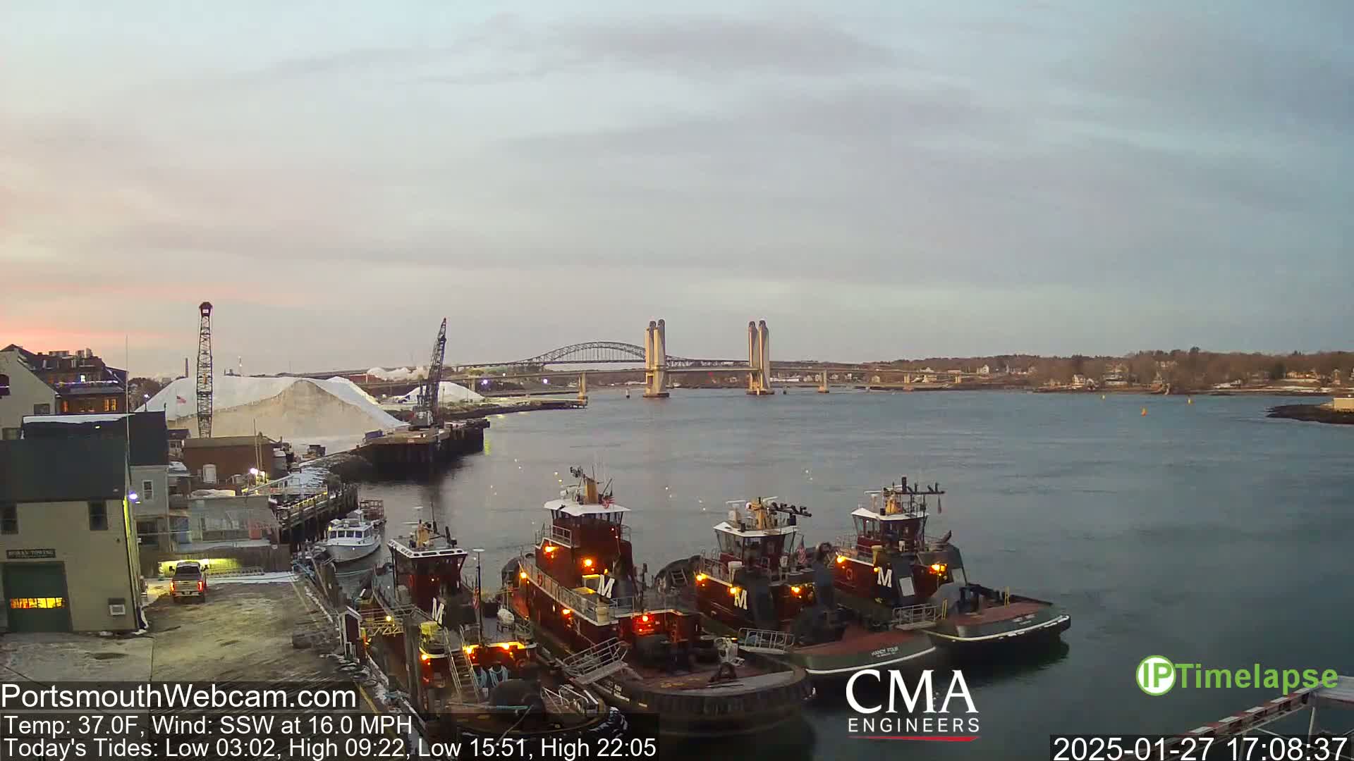 Several tugboats are moored at a dock next to a large pile of white material, with a double-lift bridge and calm water visible in the background under a pale, overcast sky.