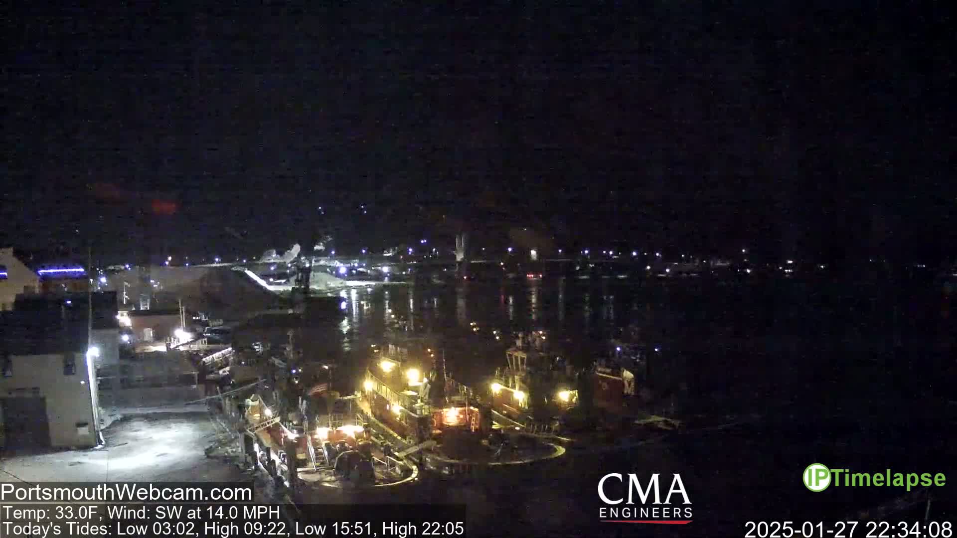 Several tugboats are illuminated at a nighttime dockside, with a calm body of water in the background under a dark sky.