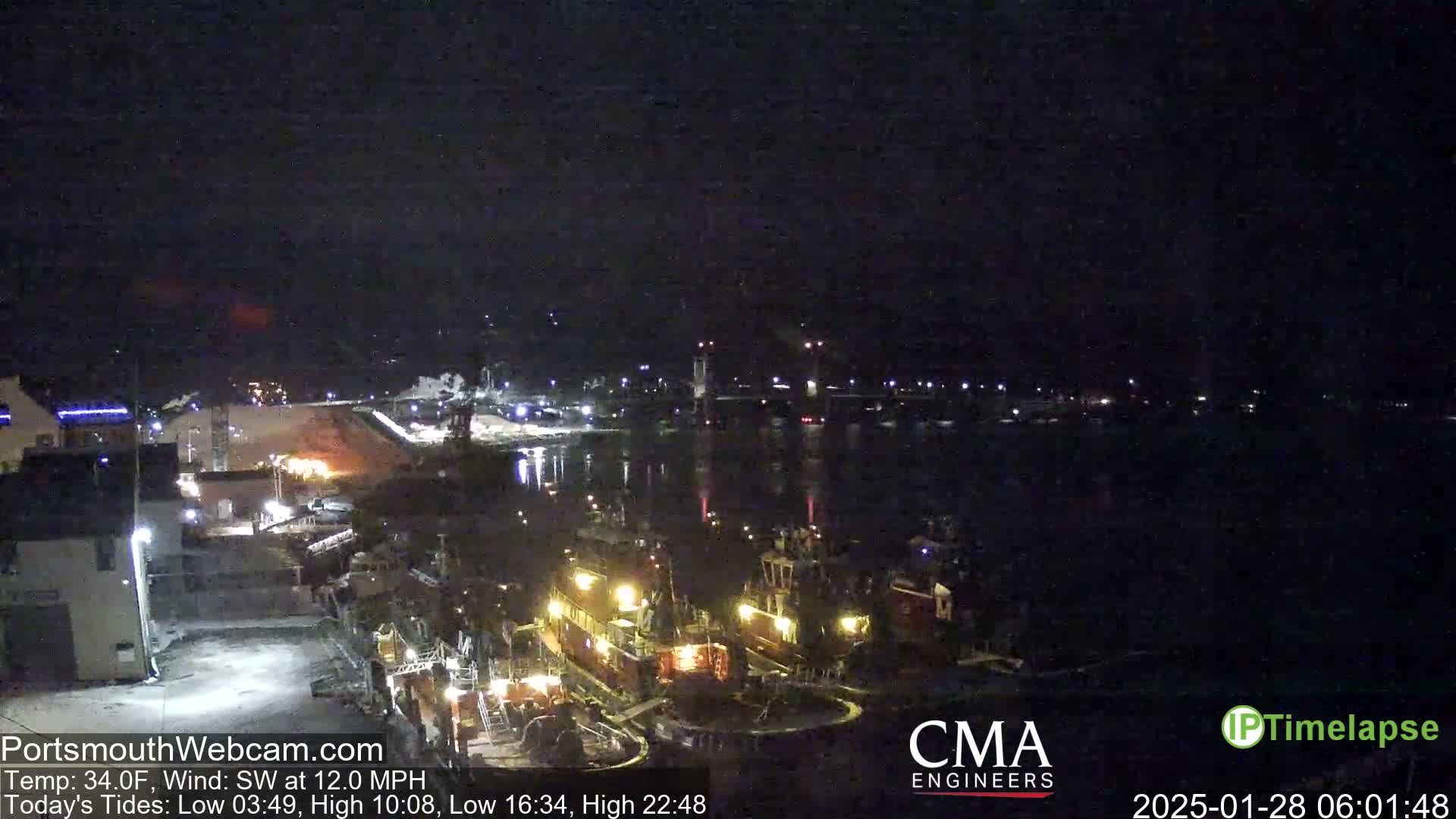 At night, under a dark sky with a southwest wind of 12 mph and a temperature of 34°F, two tugboats are moored near a waterfront industrial area with a bridge visible in the distance.