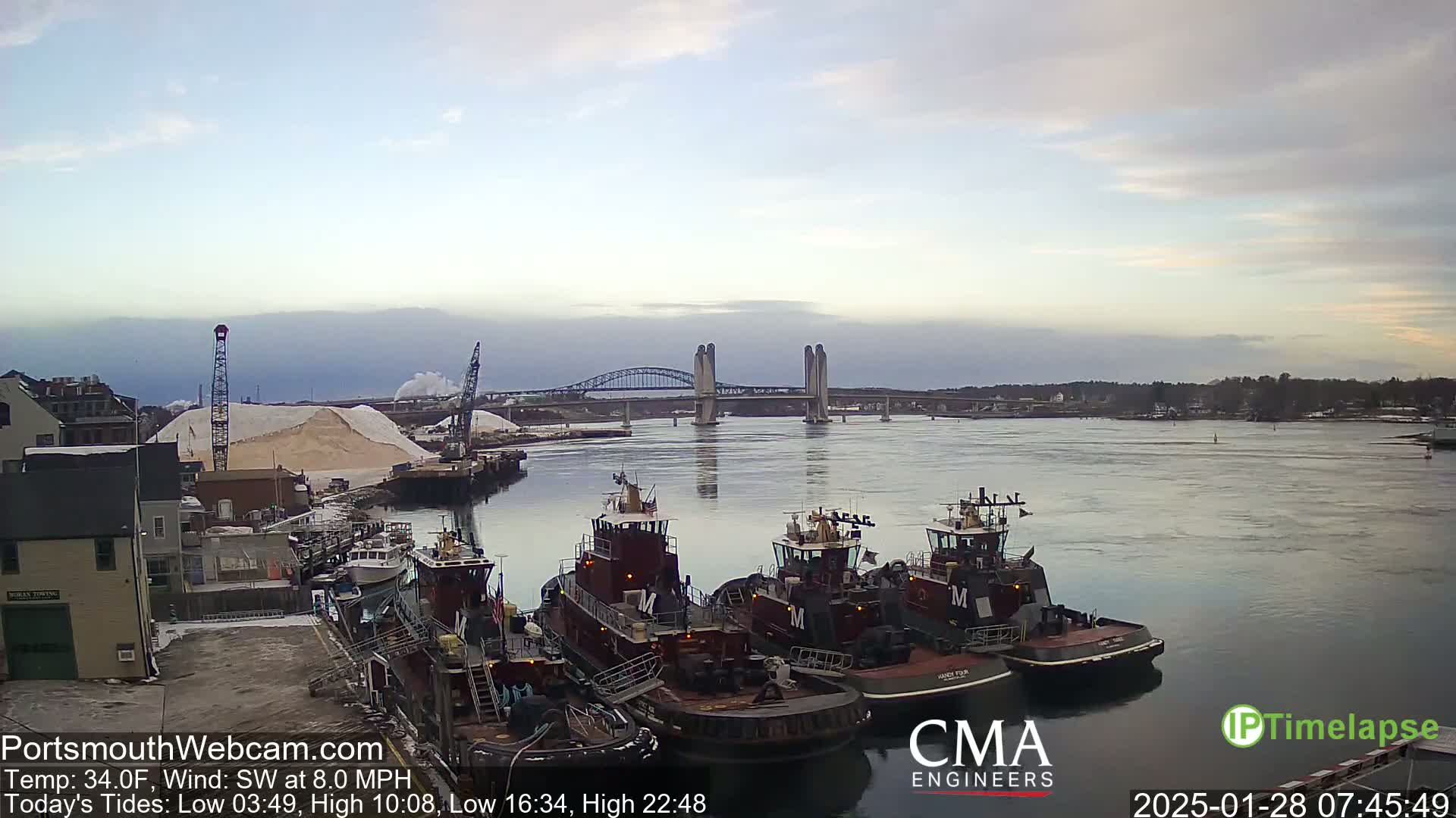 Several tugboats are moored at a dock next to a pile of sand, with a bridge spanning a calm river in the background under a cloudy sky.