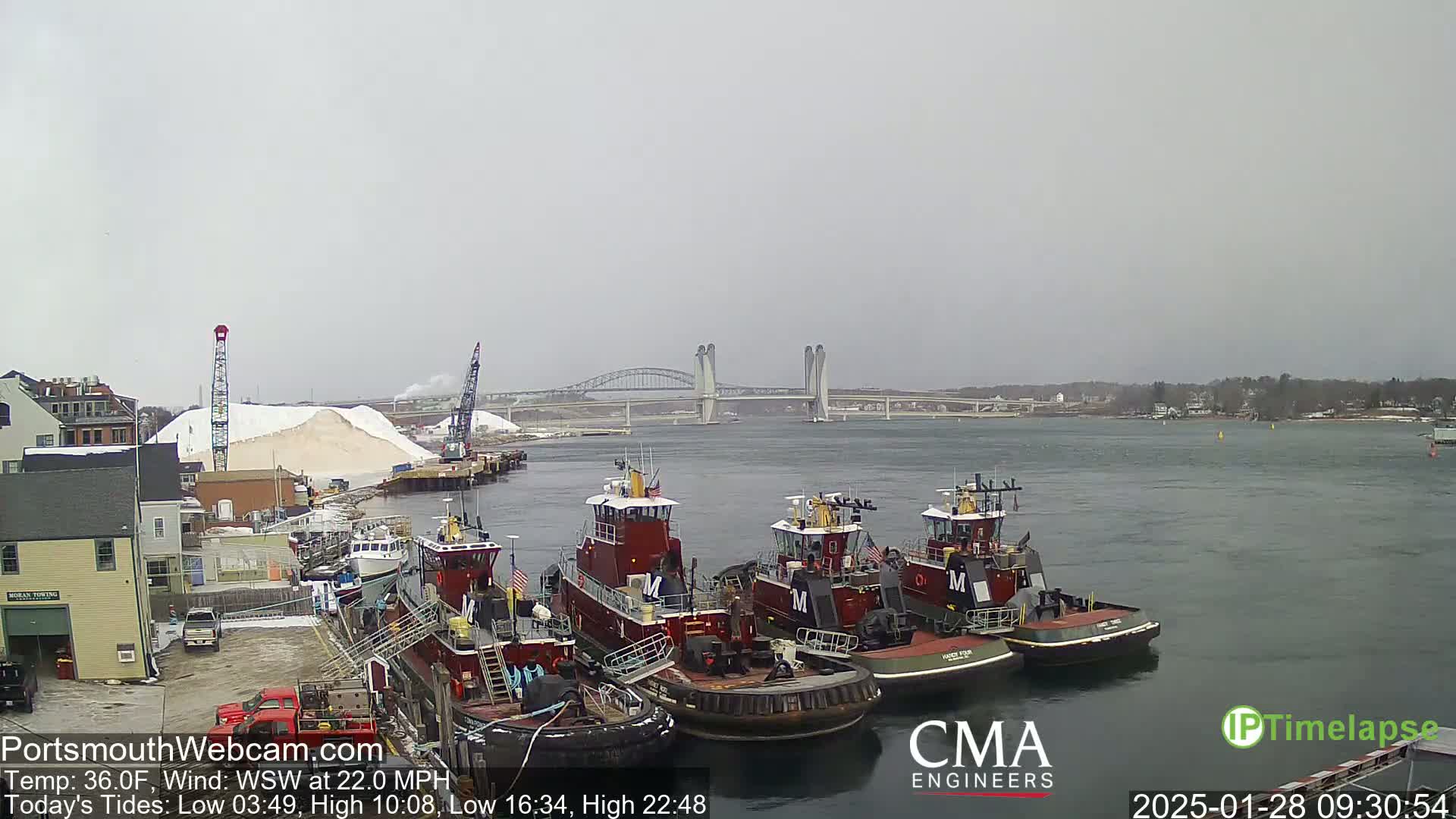 Several tugboats are docked near a large pile of salt, with a bridge visible in the distance under an overcast sky.