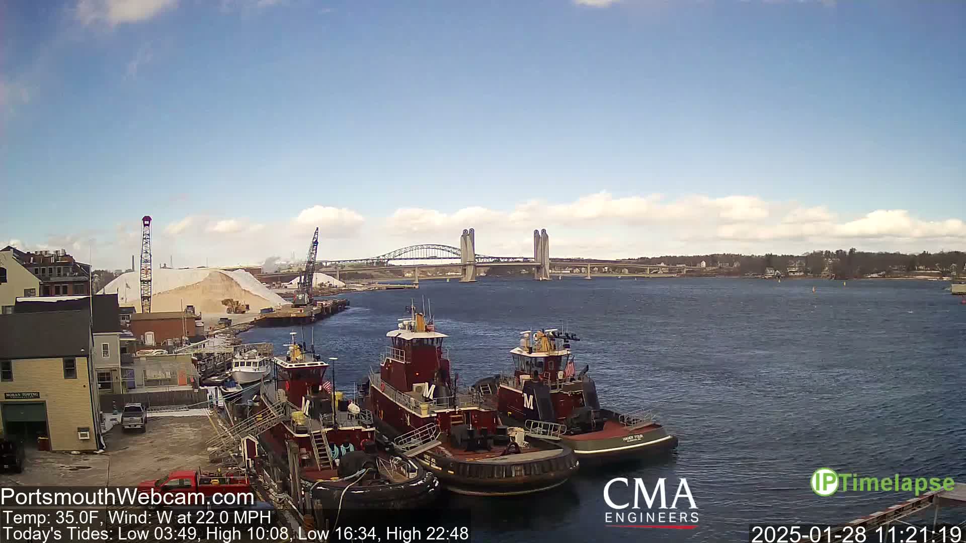 Several tugboats are moored at a waterfront dock under a partly cloudy sky, with a large pile of what appears to be salt nearby and a bridge in the distance.