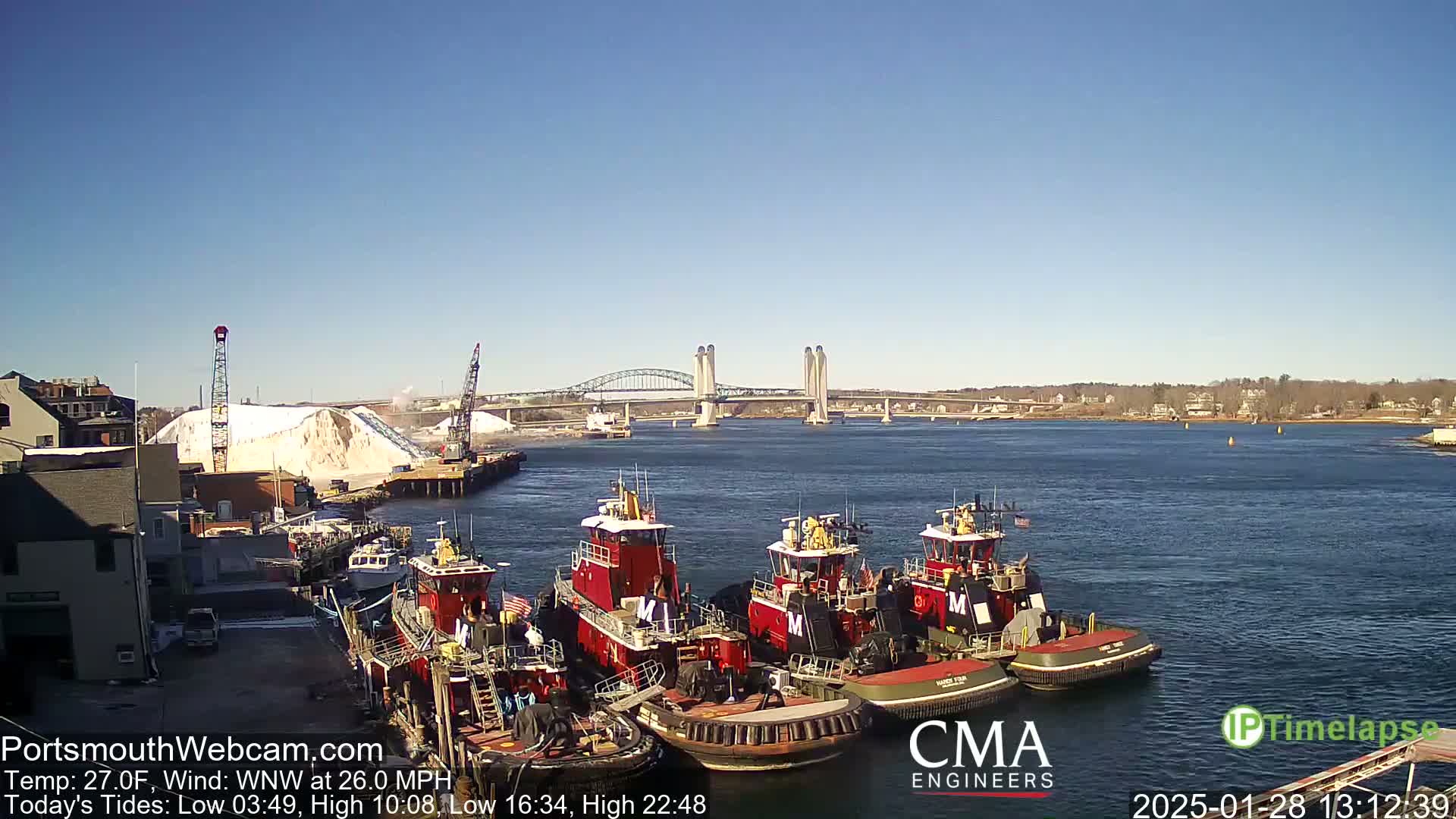 Several tugboats are moored at a dock next to a large pile of snow, with a bridge spanning a waterway in the background under a clear, sunny sky.