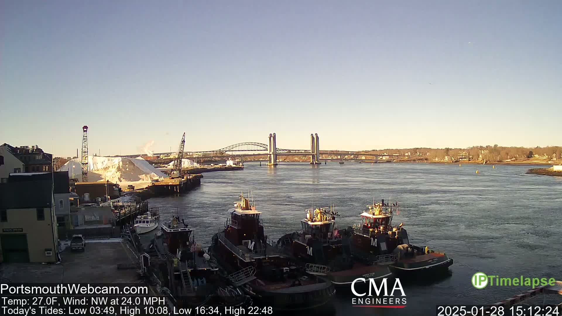 Several tugboats are moored at a waterfront dock, with a bridge and a large pile of snow visible in the distance on a clear, cold, windy day.