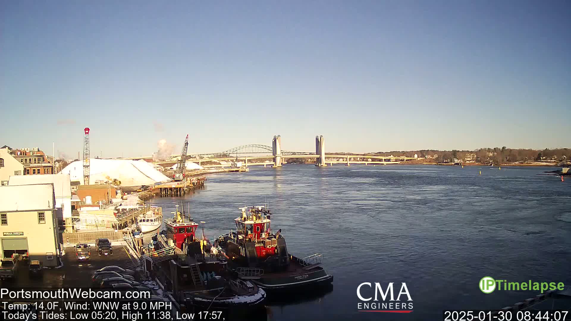 A clear, cold day reveals two tugboats moored near a waterfront, a large pile of white material, and a lift bridge spanning a wide waterway.