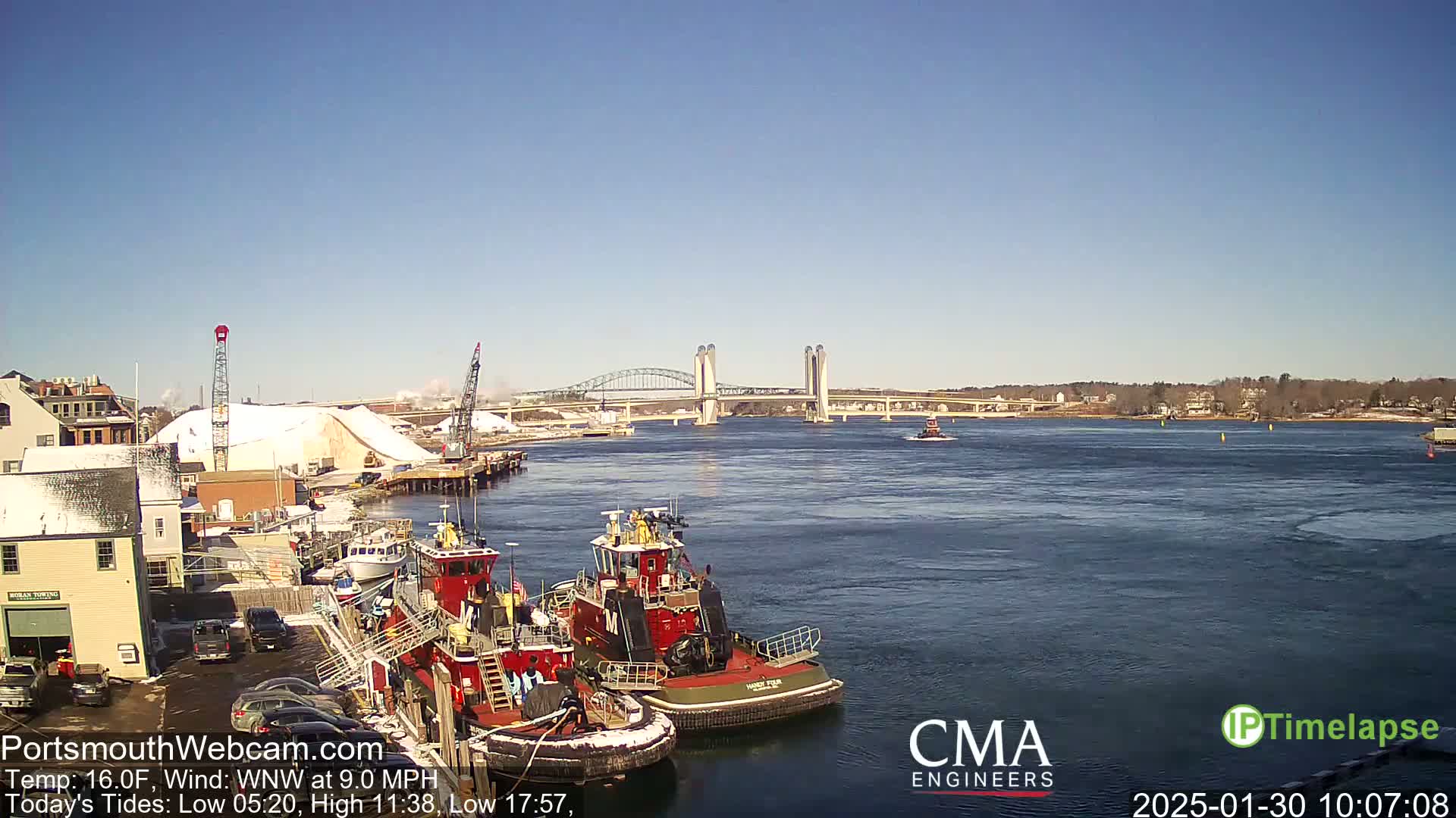 A clear, cold day shows two tugboats moored near a waterfront with a snow-covered pile of material, cranes, and a lift bridge in the background.