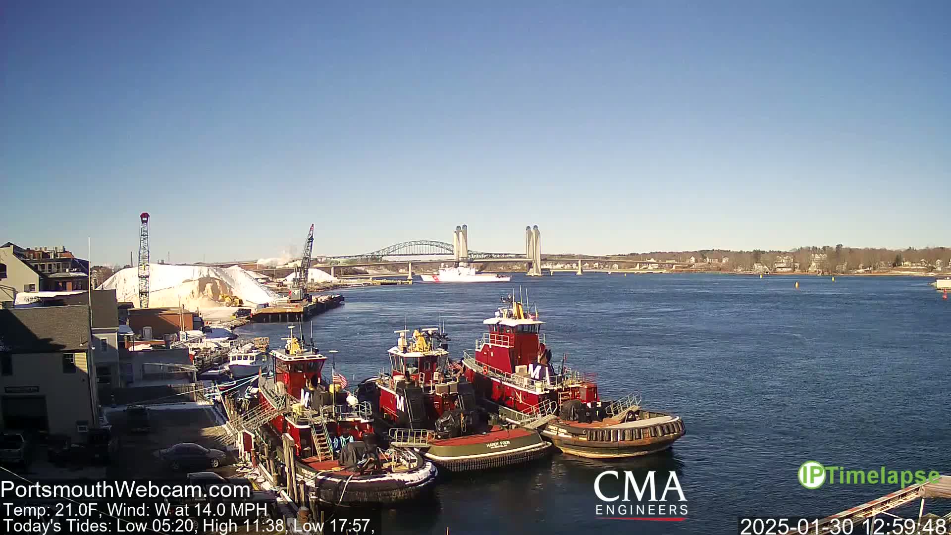 Several tugboats are docked at a harbor, with a bridge and a large pile of snow visible in the background under a clear, sunny sky.