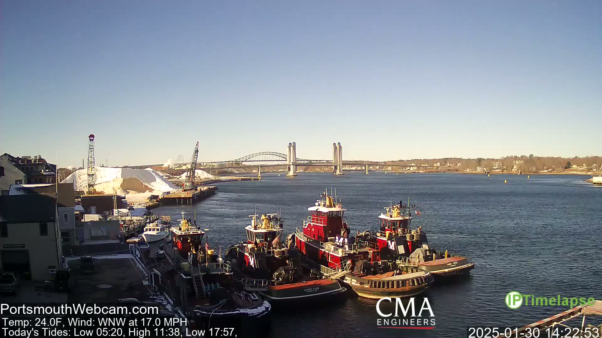 Several tugboats are moored at a dock next to a large pile of snow, with a bridge visible in the distance under a clear, sunny sky.