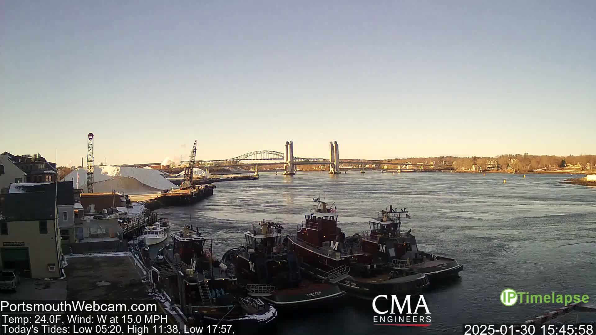 Several tugboats are docked near a large pile of white material, with a bridge in the background under a clear, cold, and windy sky.