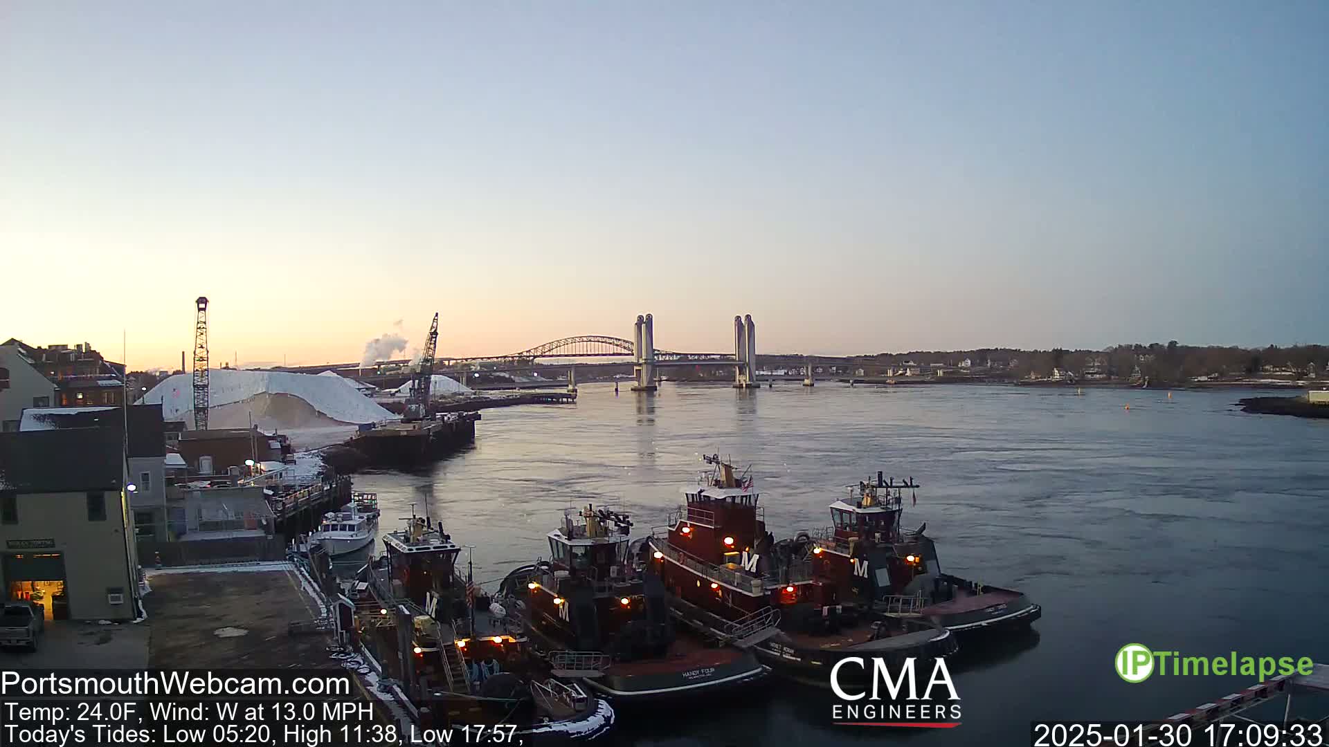 Several tugboats are moored at a dock near a bridge at sunset under a clear sky with a light west wind.