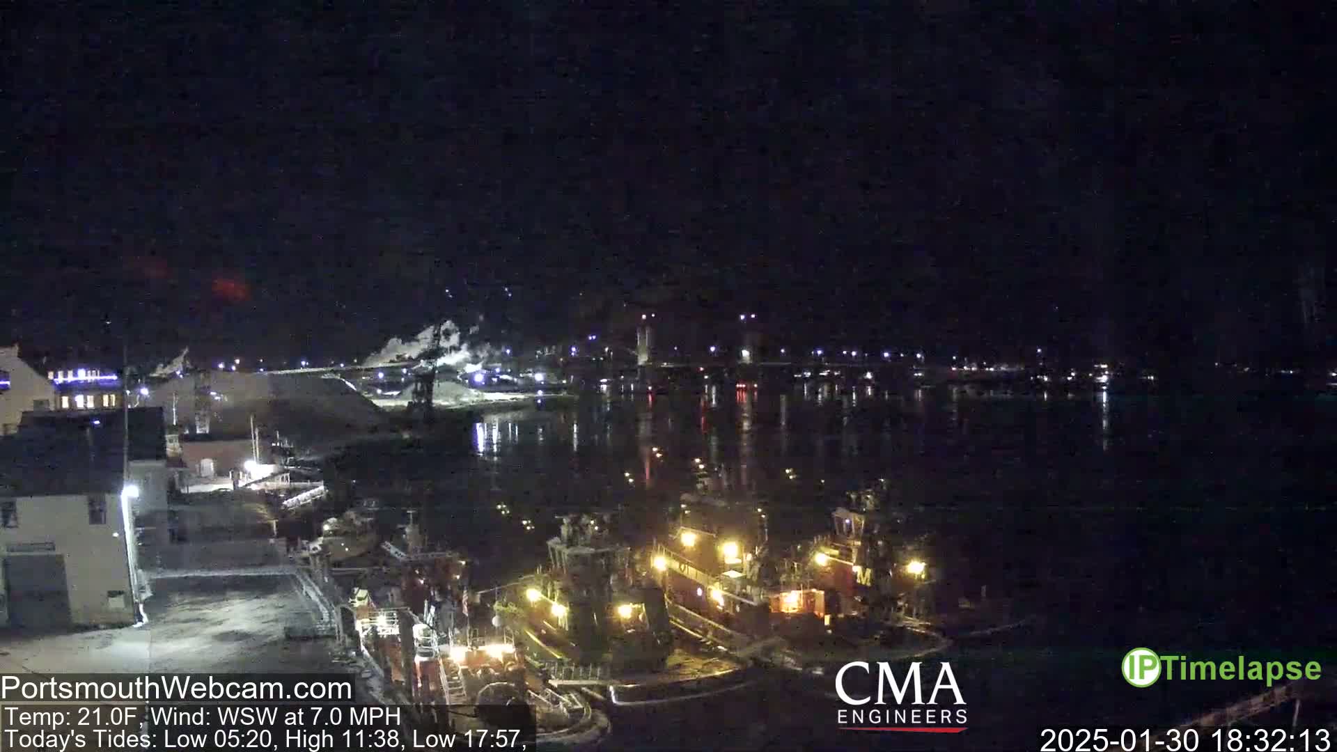 At night, two tugboats are docked near a shoreline with industrial buildings in the background, under a dark, clear sky.