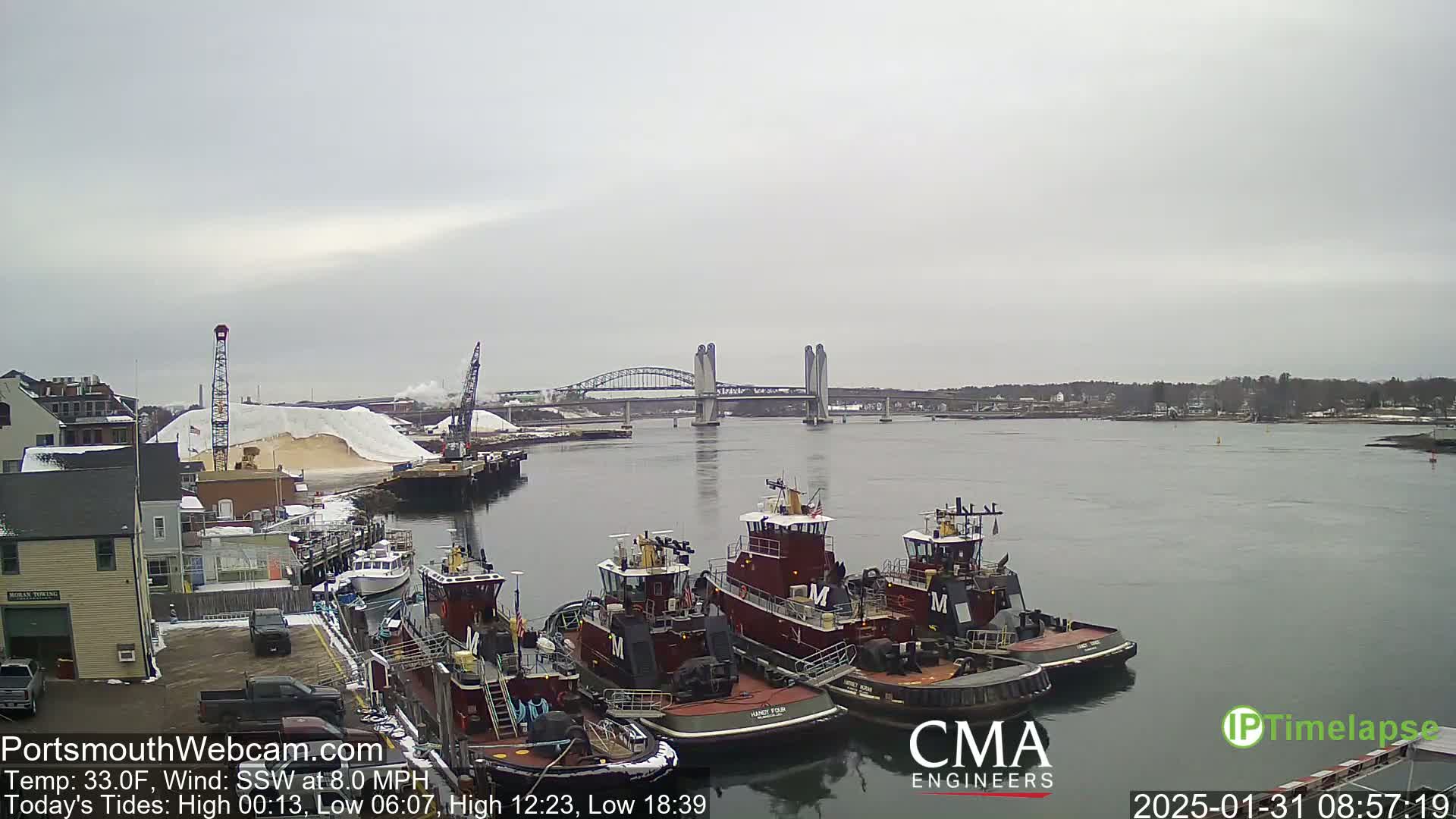 Several tugboats are moored at a dock next to a large pile of sand, with a bridge spanning a calm river in the background under an overcast sky.