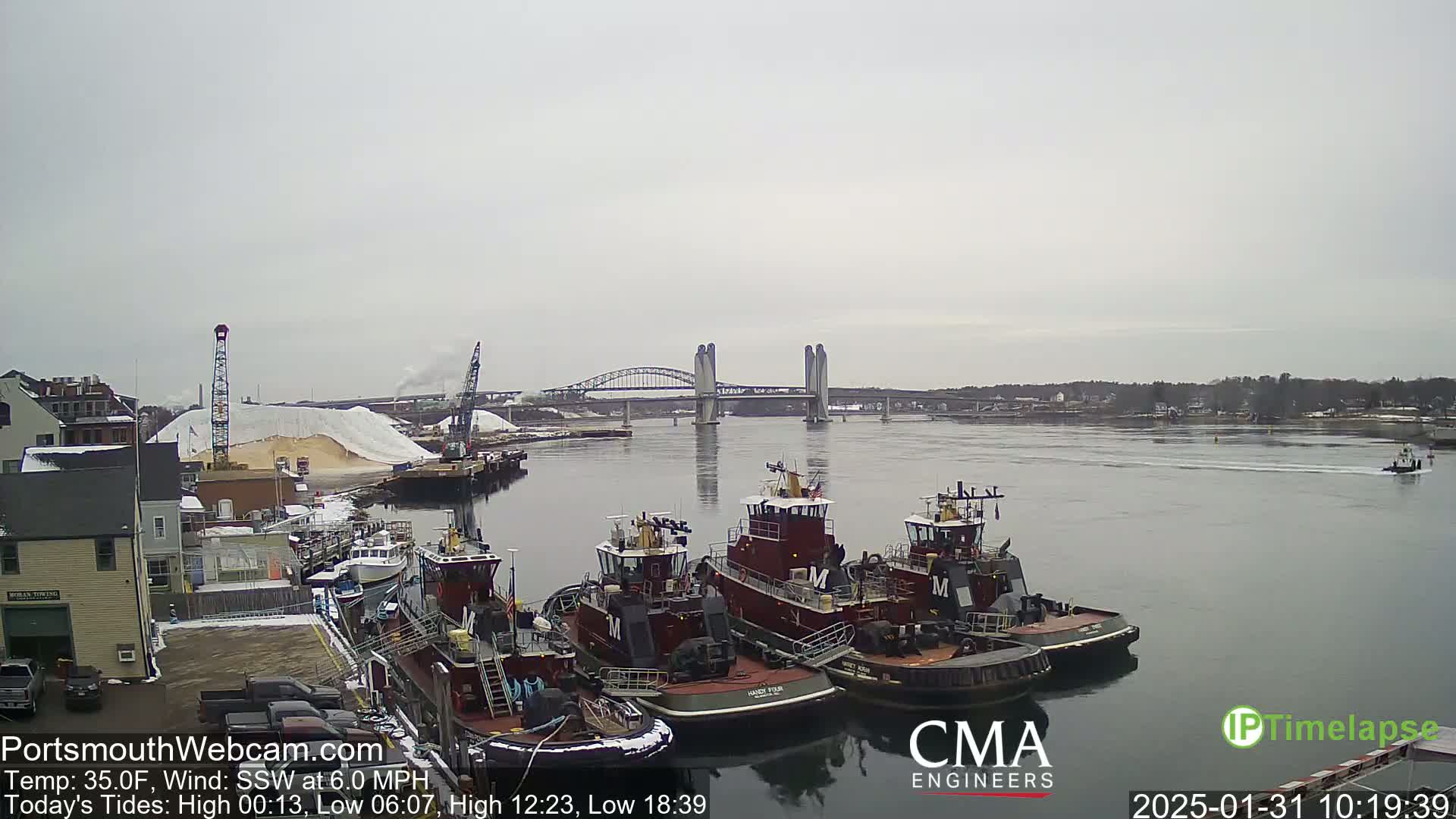 Several tugboats are moored at a dock near a bridge on a cloudy day, with a large pile of light-colored material visible in the background.