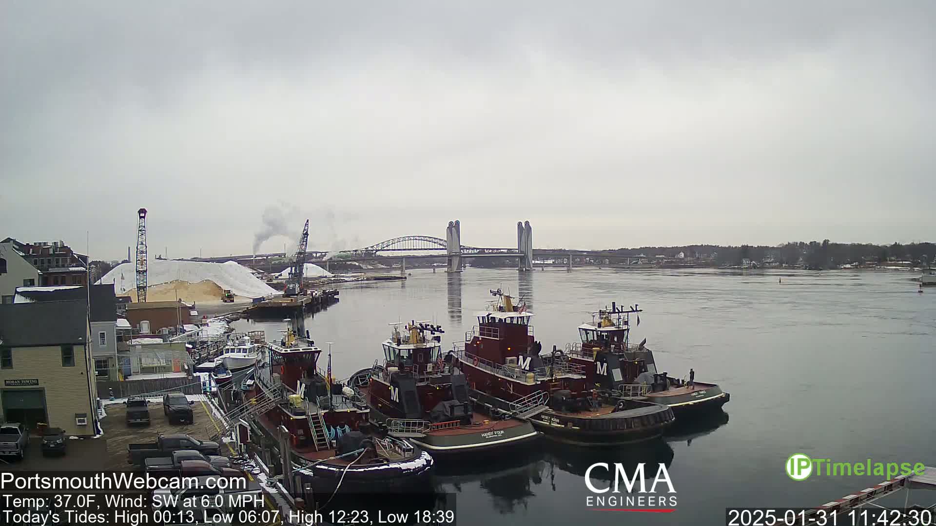 Three tugboats are docked at a waterfront, with a large pile of white material and a bridge in the background under an overcast sky.