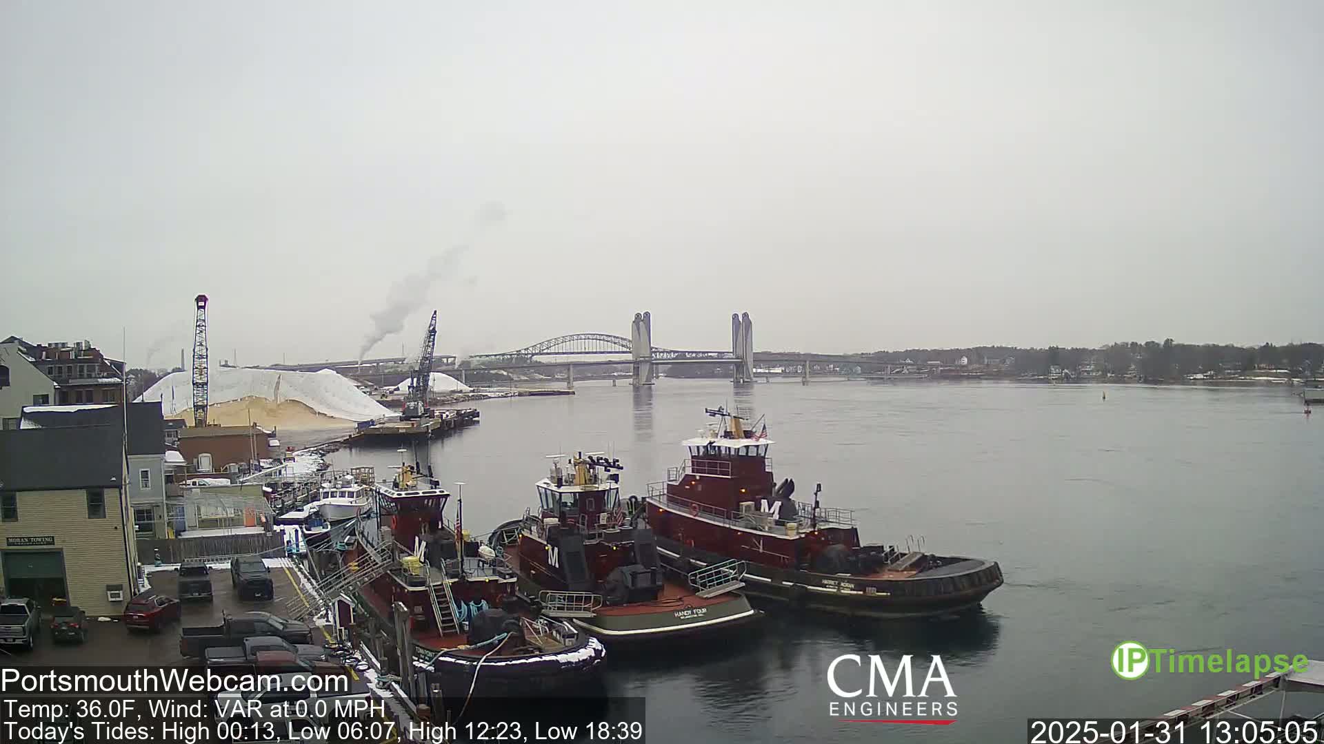 Three tugboats are moored near a large pile of tan material next to a bridge over a calm, gray river under an overcast sky.