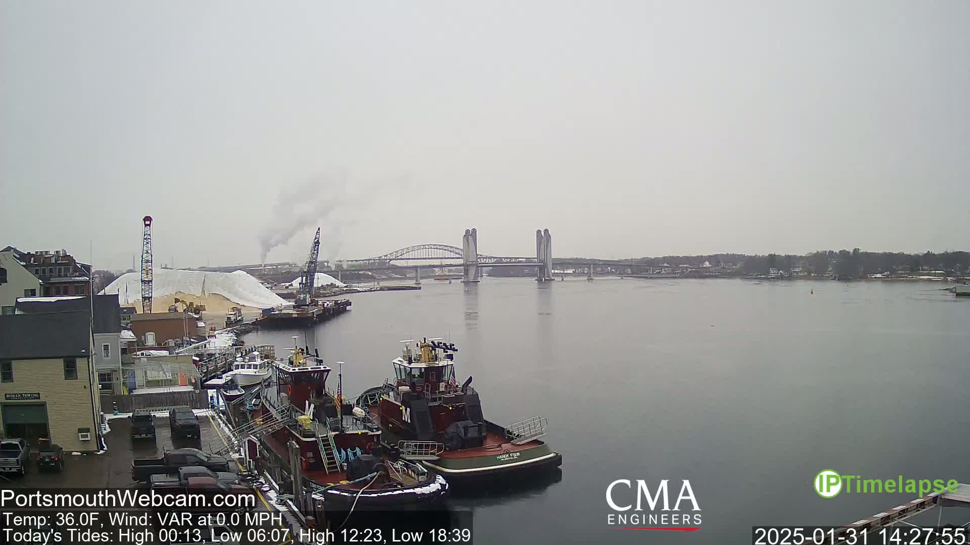 A harbor scene on an overcast day shows tugboats docked near a large pile of sand, with a bridge in the distance.