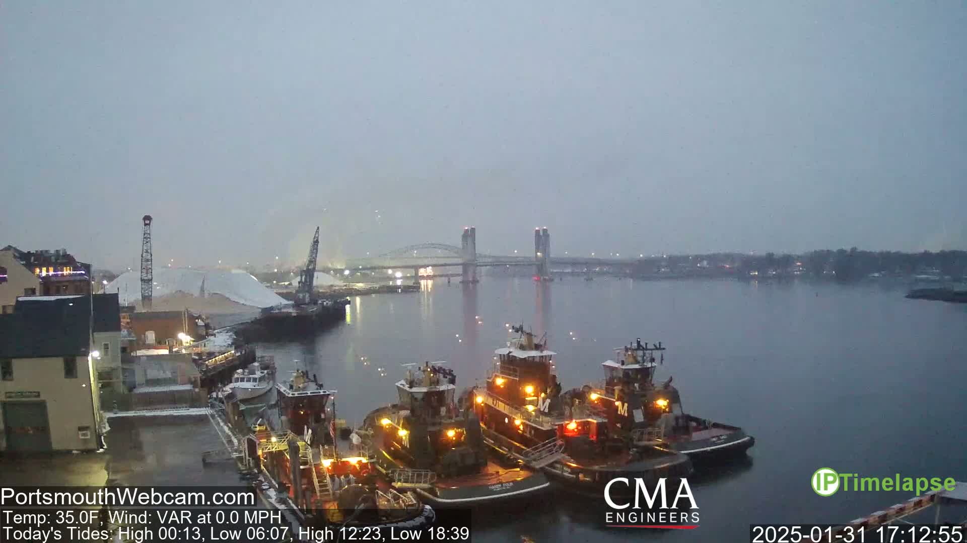 Three tugboats are moored at a dock near a bridge in overcast conditions.