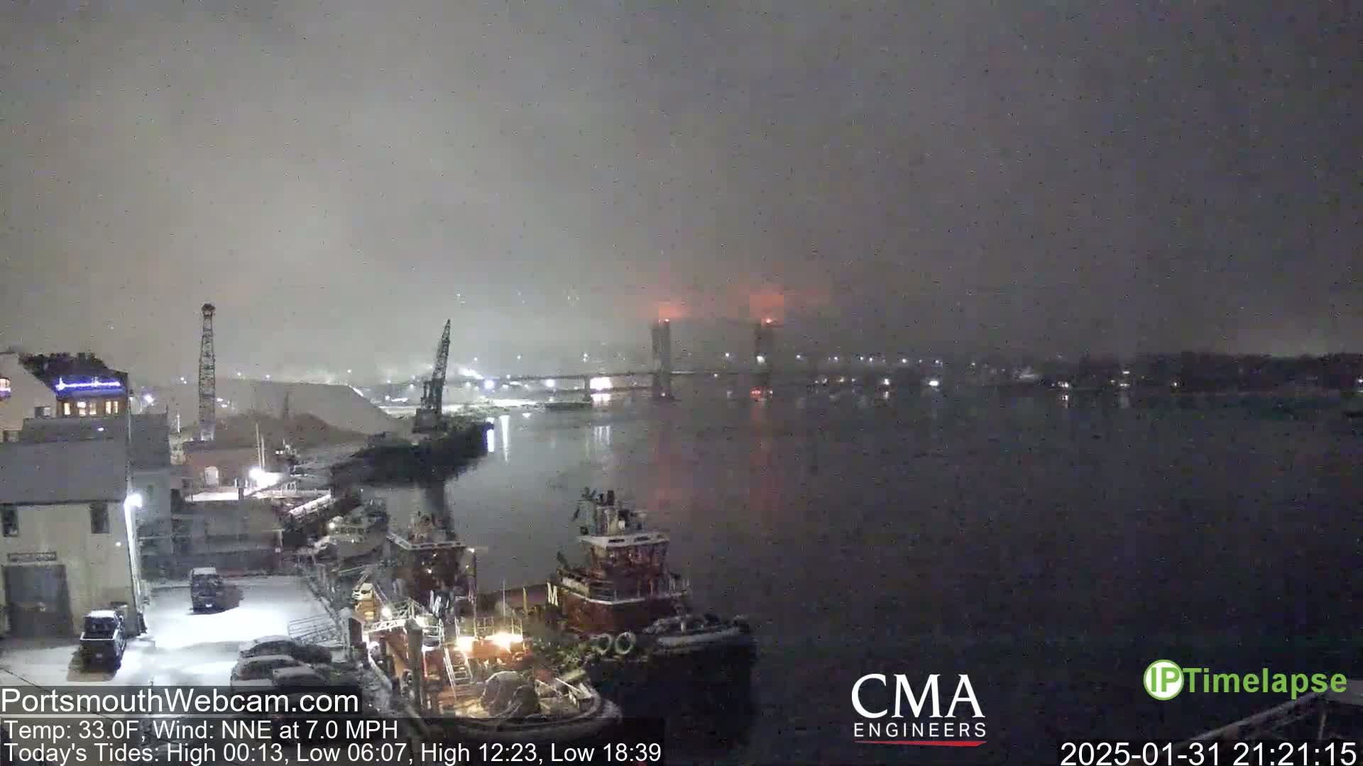 A nighttime view of a snow-covered harbor with tugboats and a bridge in the distance under foggy conditions.