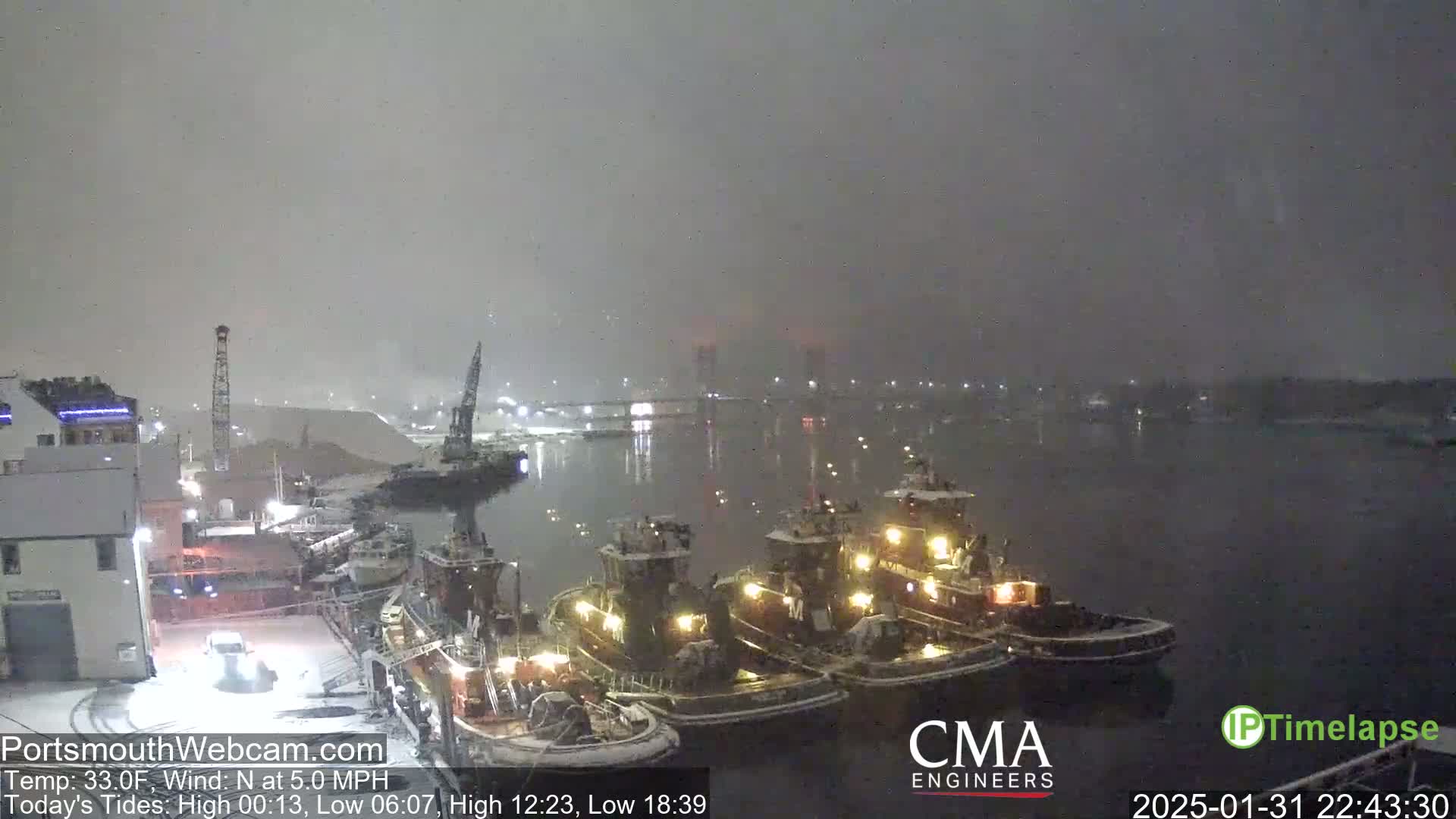 Three tugboats are docked in a snow-covered harbor at night, with a bridge and other lights visible in the distance under a heavy snowfall.