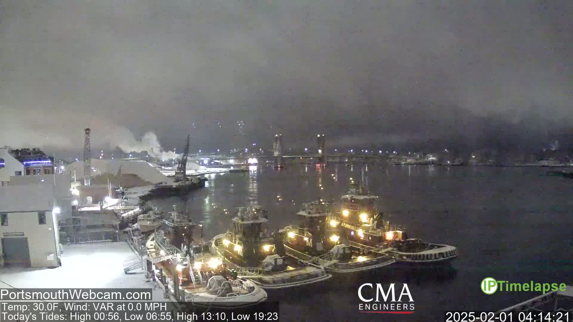 Three tugboats are moored at a snow-covered dock at night under a dark, cloudy sky.