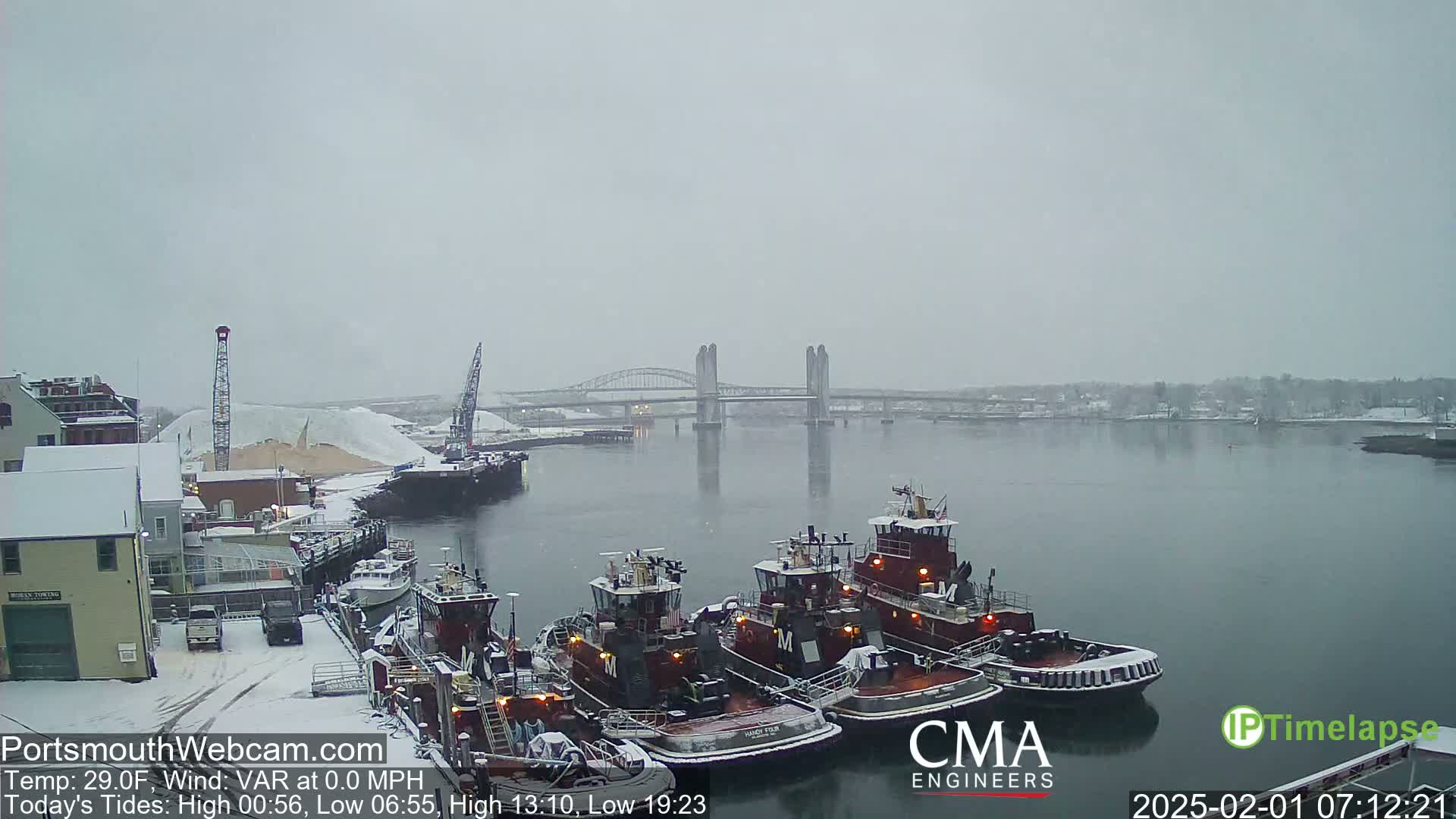 Several tugboats are docked at a snowy waterfront, with a bridge spanning a calm river under a gray, snowy sky.