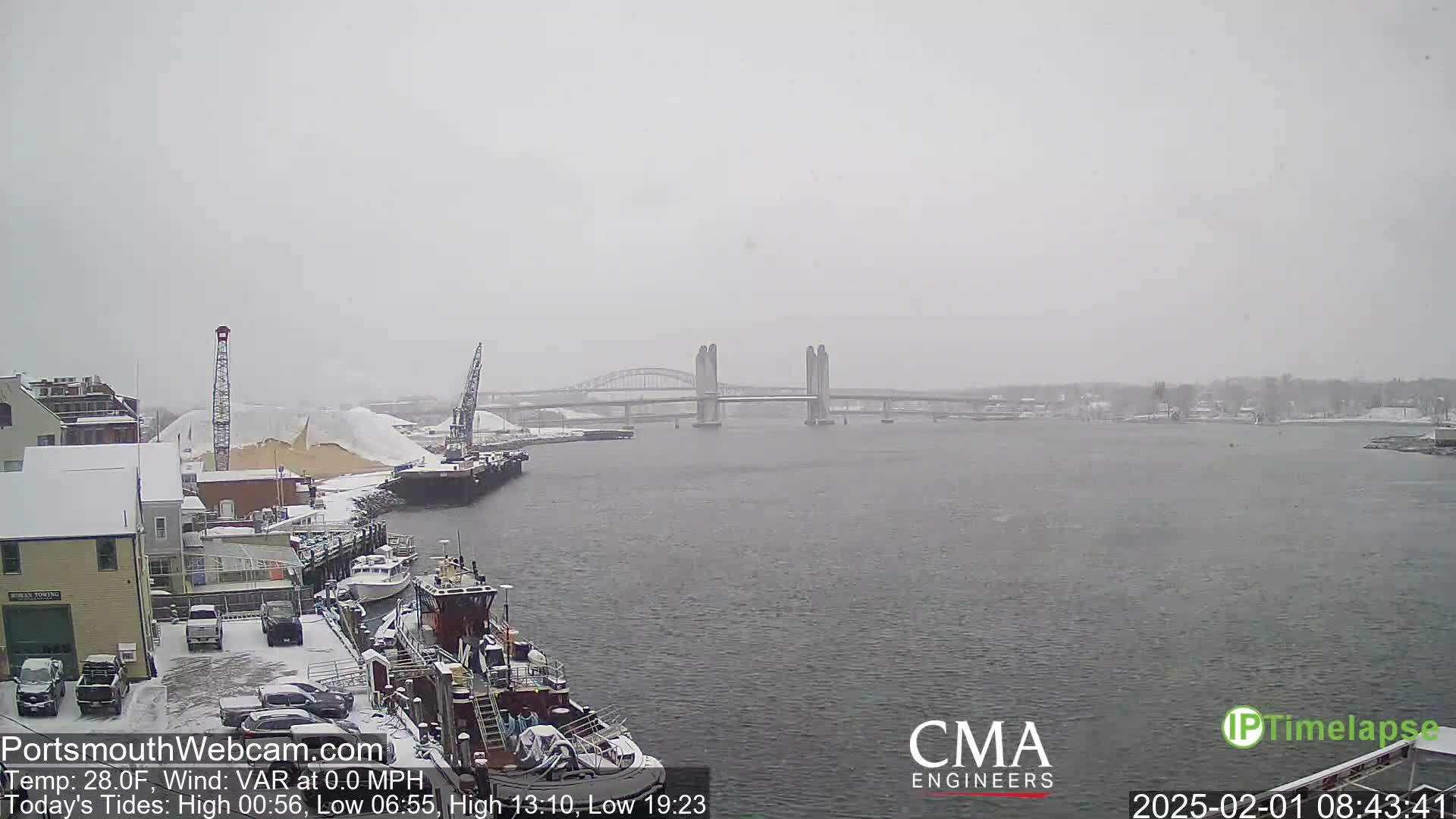 A snow-covered harbor scene shows a lift bridge over a waterway, with a tugboat and other vessels docked nearby, under a light snowfall.