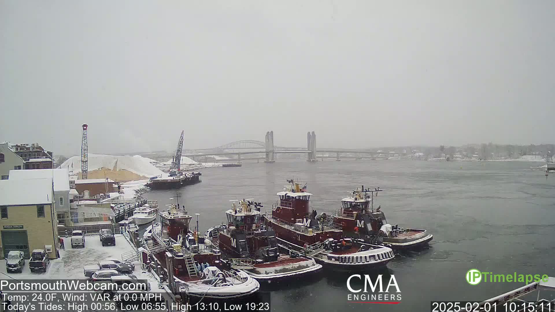 Several tugboats are docked at a snowy waterfront, with a bridge visible across a body of water in the background under a lightly snowing sky.