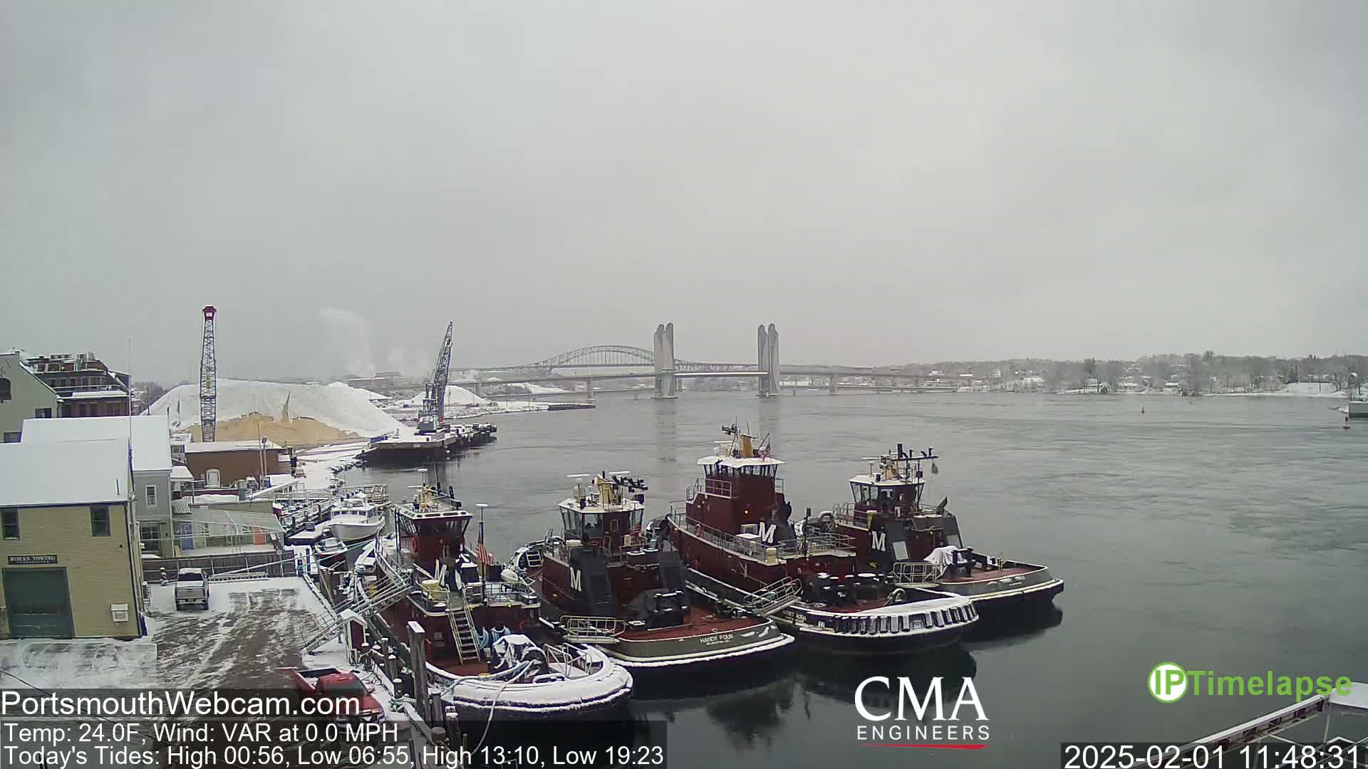 A snowy waterfront scene shows several tugboats docked near a snow-covered industrial area, with a bridge visible in the background under an overcast sky.