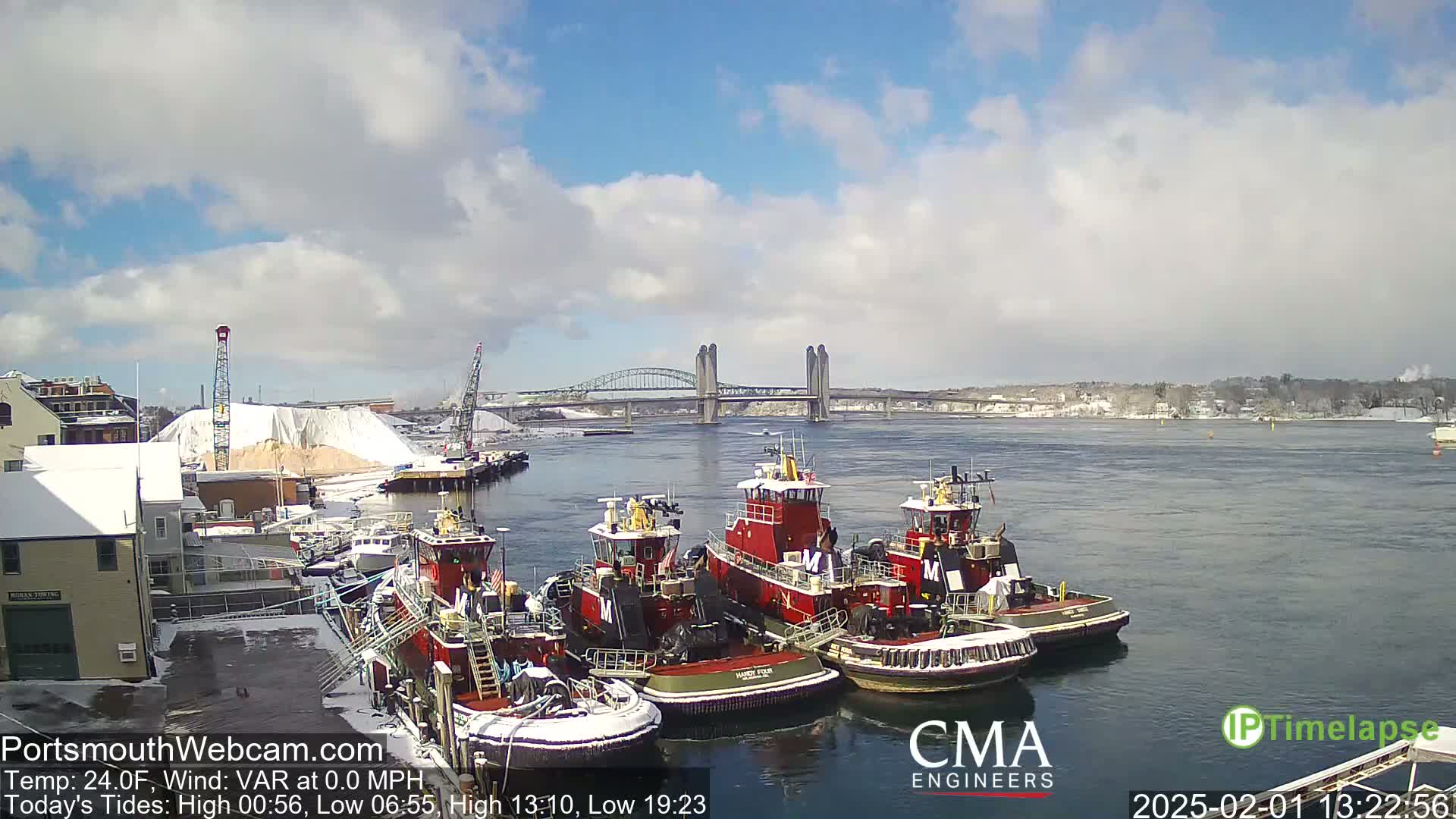 Several tugboats are docked at a snow-covered waterfront, with a bridge visible across a calm river under a partly cloudy sky.