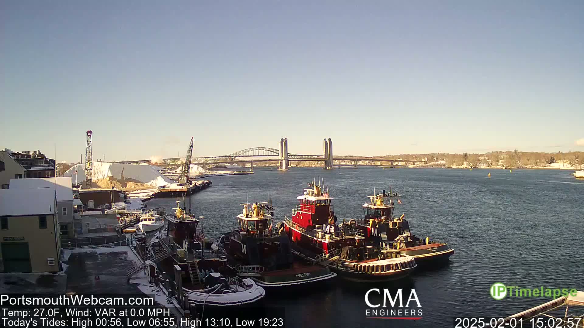 Several tugboats are docked at a waterfront, with a bridge spanning a calm body of water under a clear, sunny sky; snow is visible on the ground and nearby buildings.